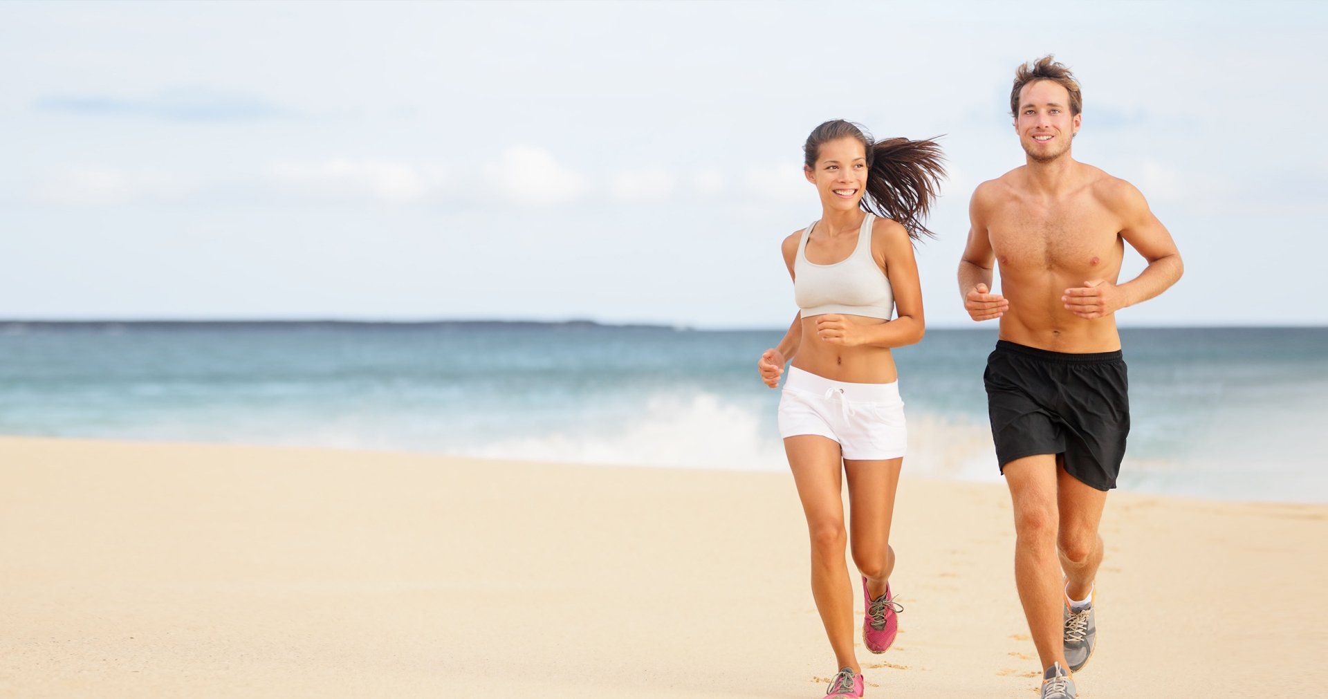 Woman and man running on a beach, both smiling. Ocean in background, sunny day.