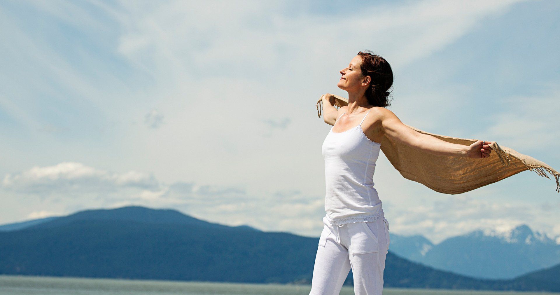 A person in white clothes stands outdoors with arms outstretched, holding a tan scarf blowing in the wind against mountains.