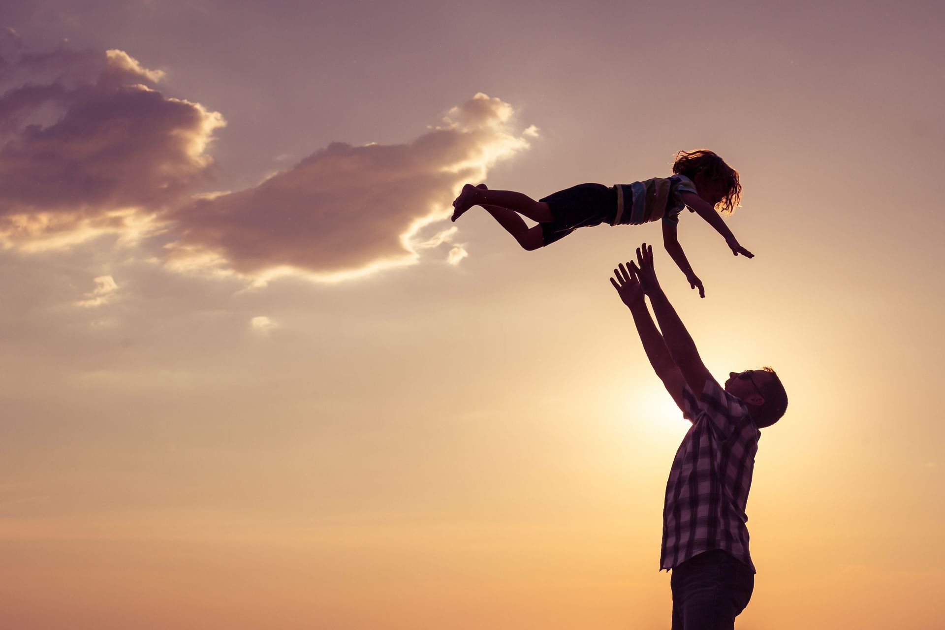 Man throwing child up into the air in front of a sunset.