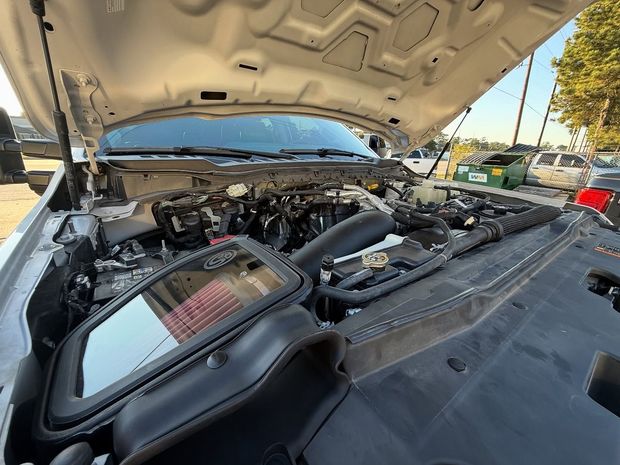 Car engine with the hood open, revealing components like the air filter and engine block in an outdoor setting.