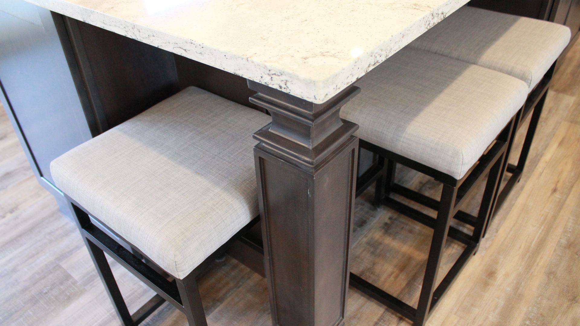Close-up of kitchen island with three gray upholstered stools. Brown island support. Light countertops and floor.
