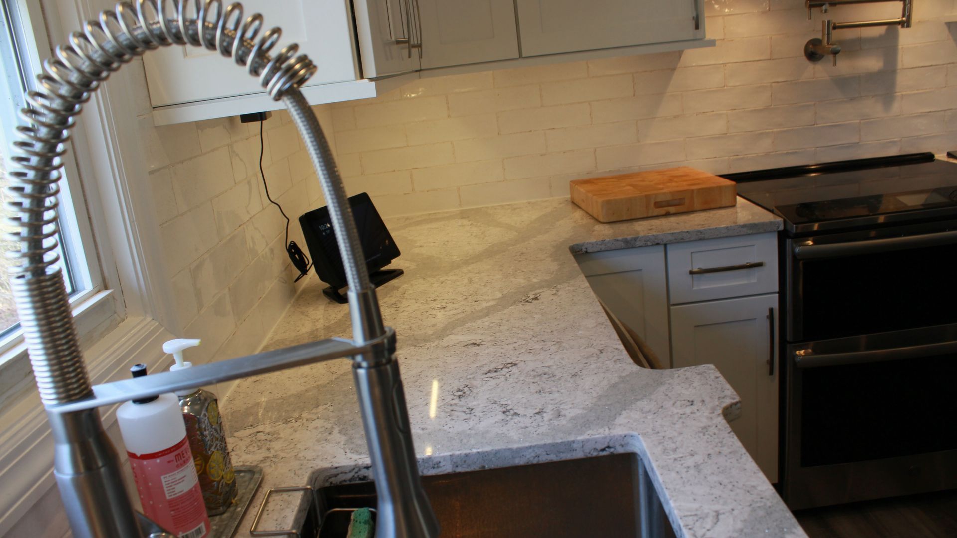 Kitchen with stainless steel faucet, white cabinets, speckled countertop, cutting board, and oven.