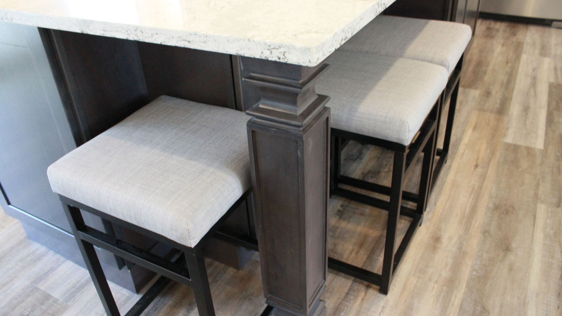 Kitchen island with two gray upholstered stools; dark wood cabinetry and light flooring.