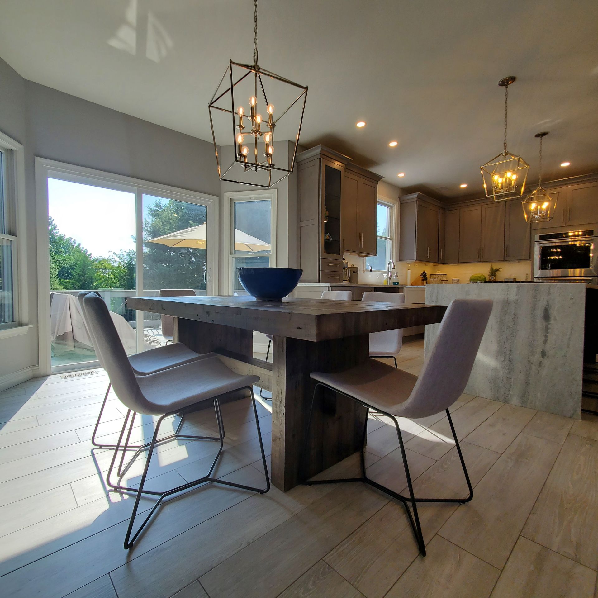 Dining room with wooden table, chairs, and overhead light fixtures. Kitchen in background.