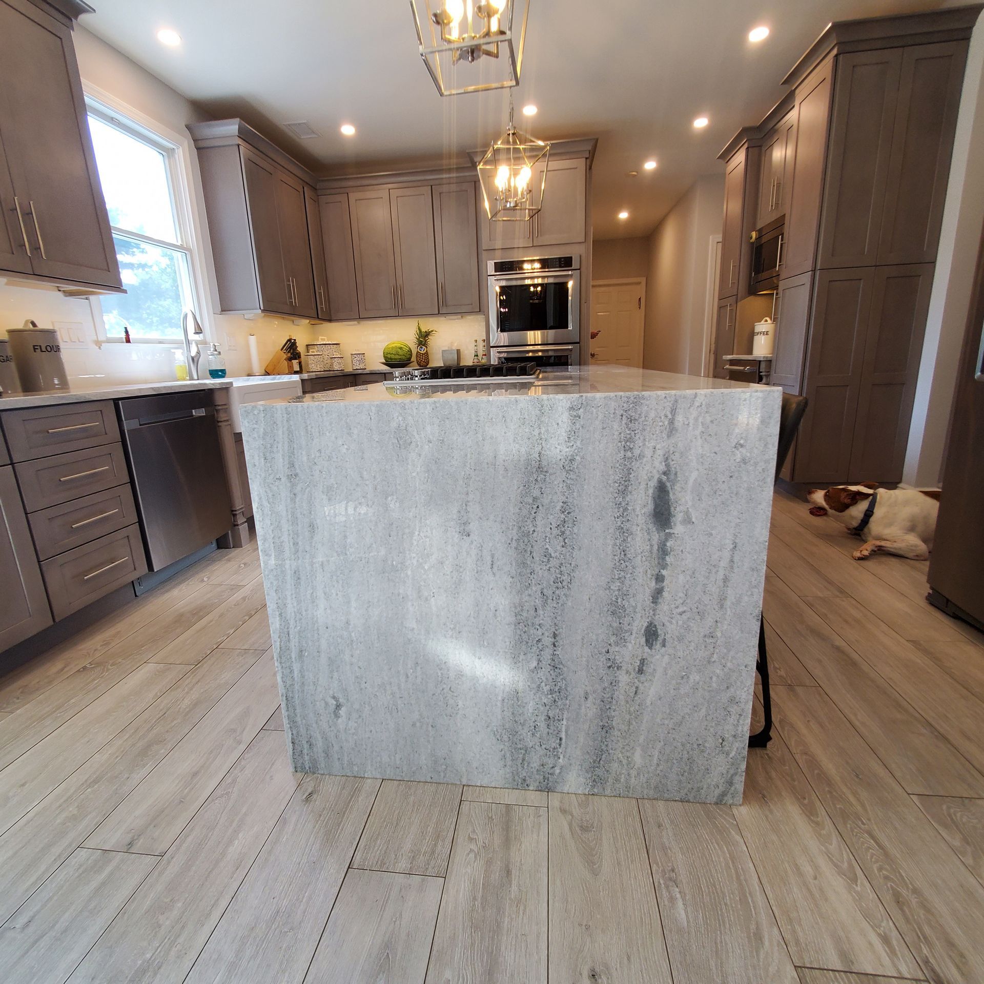 A modern kitchen with grey cabinets, a marble island, and a dog lying on the floor.