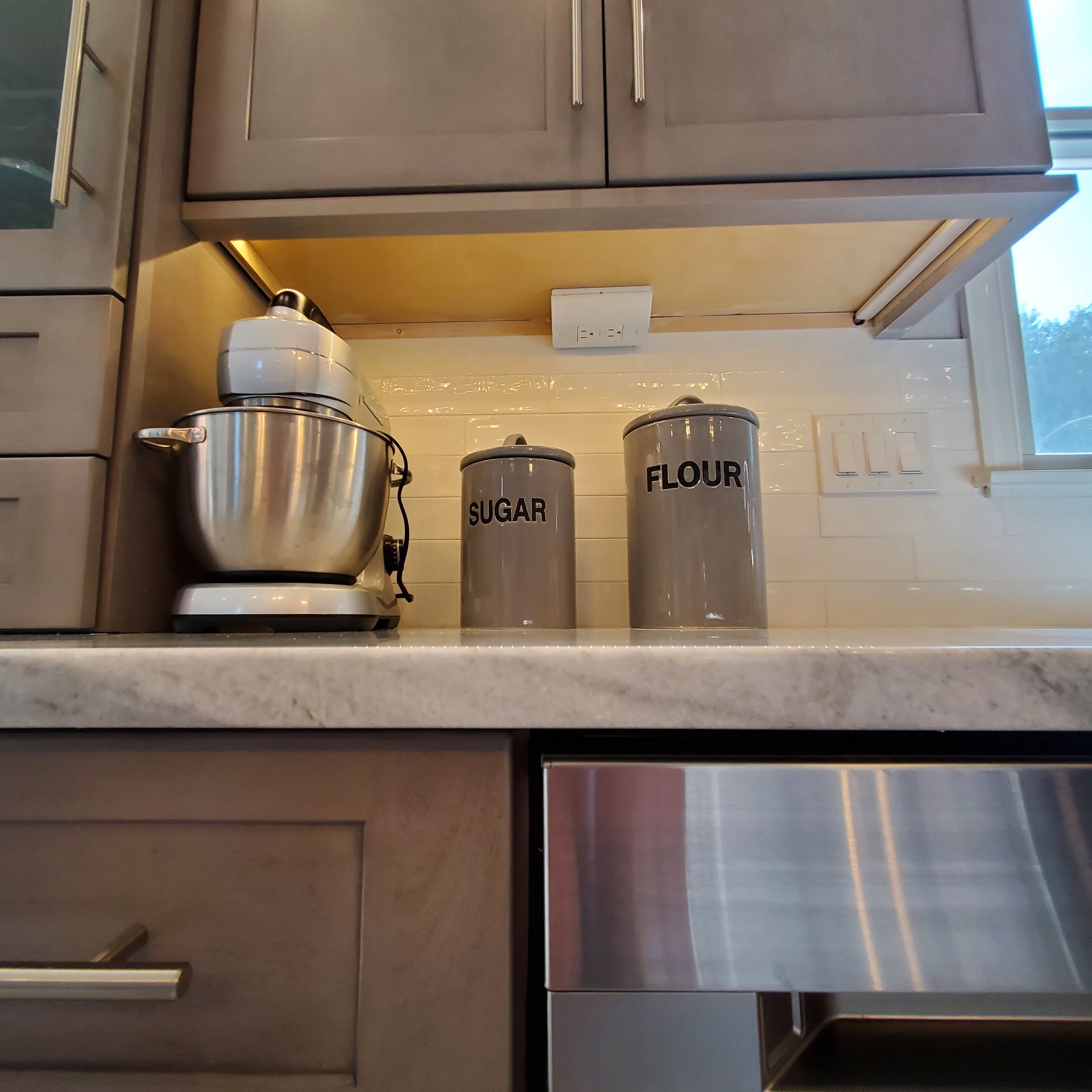 A kitchen countertop with a mixer, sugar and flour canisters, and a stainless steel appliance.