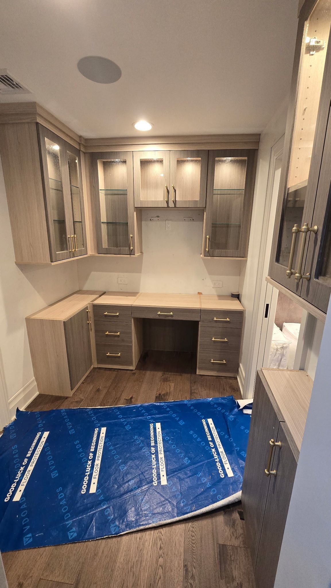 Built-in office with light-colored cabinets, desk, and overhead storage. Blue protective covering on the floor.