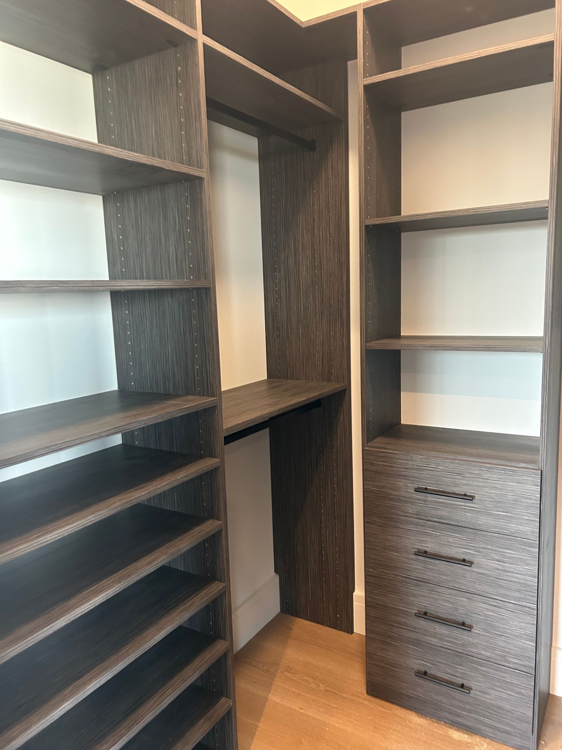 Built-in dark wood closet with shelves, drawers, and a hanging rod, set against white walls and light wood floor.