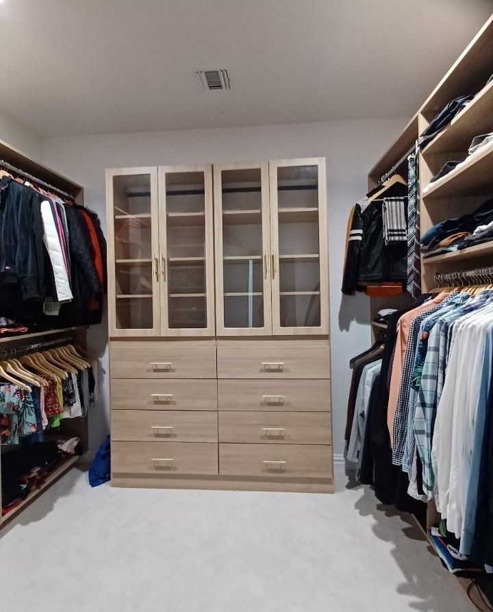 Walk-in closet with light wood cabinets and glass-doored storage, clothes hanging on either side, carpeted floor.
