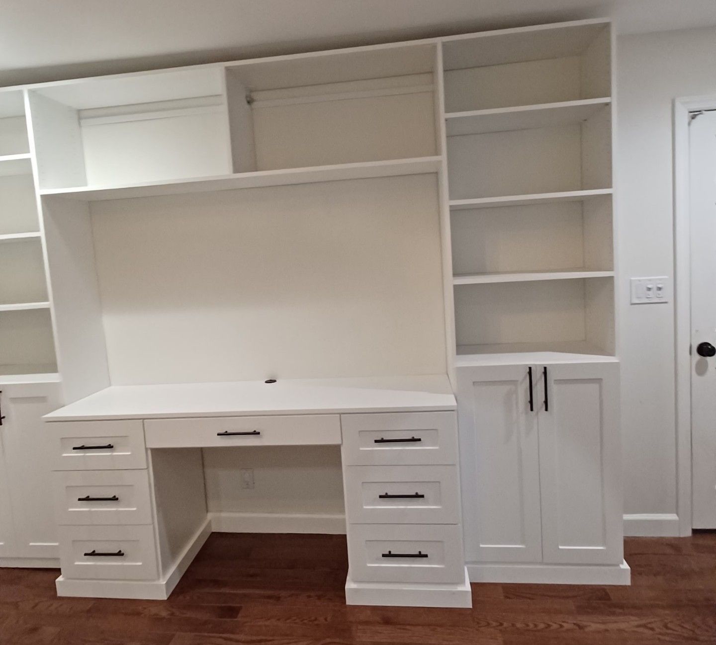 White built-in desk and shelving unit with drawers, cabinets, and bookshelves, against a light-colored wall.
