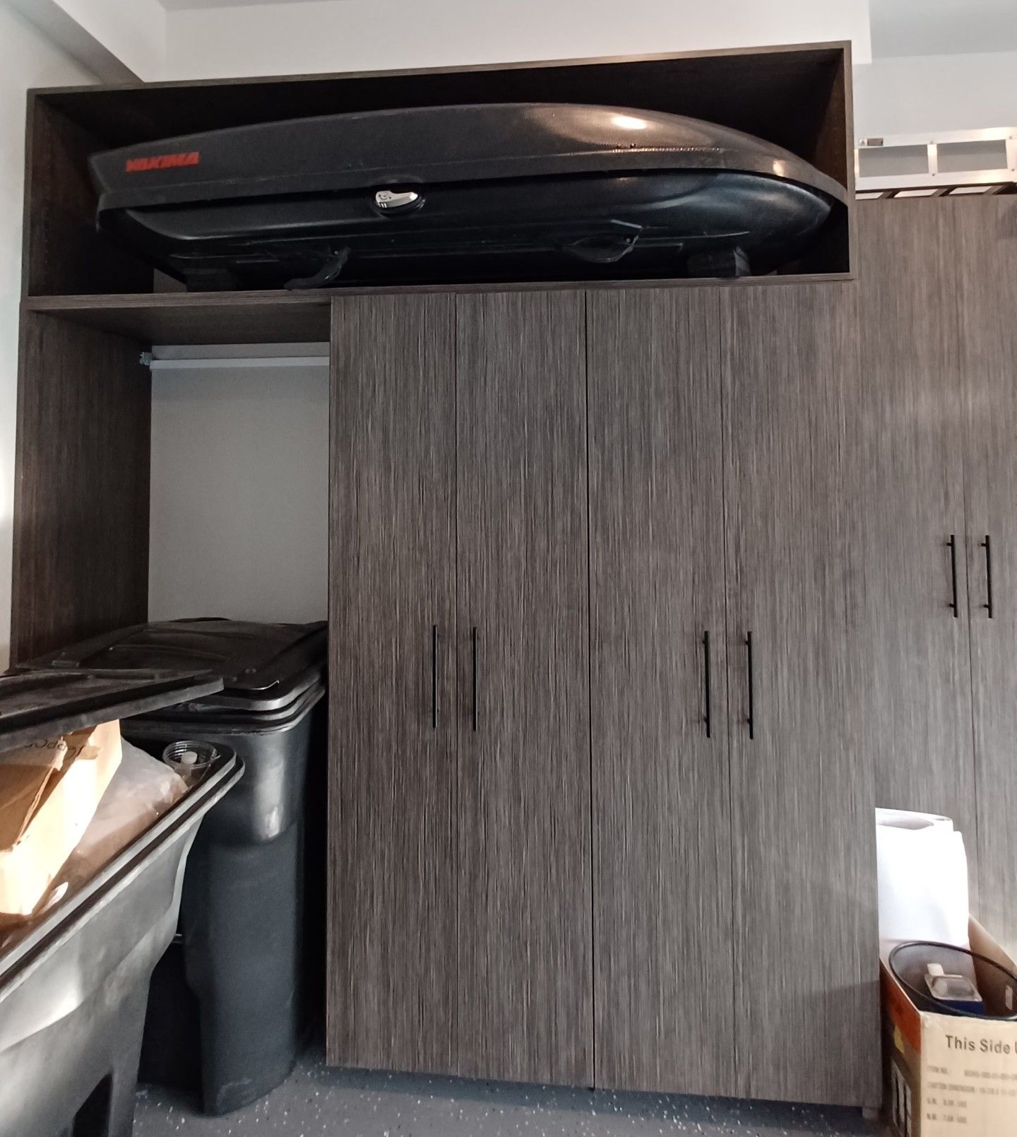 Dark wood garage storage with a cargo carrier on top. Black trash can and items on the left.
