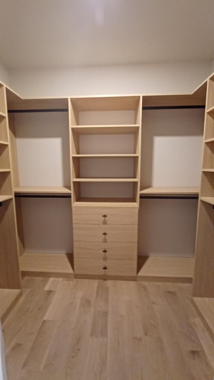 Light wood walk-in closet with shelves, drawers, and hanging rods. Beige walls, wood floor.