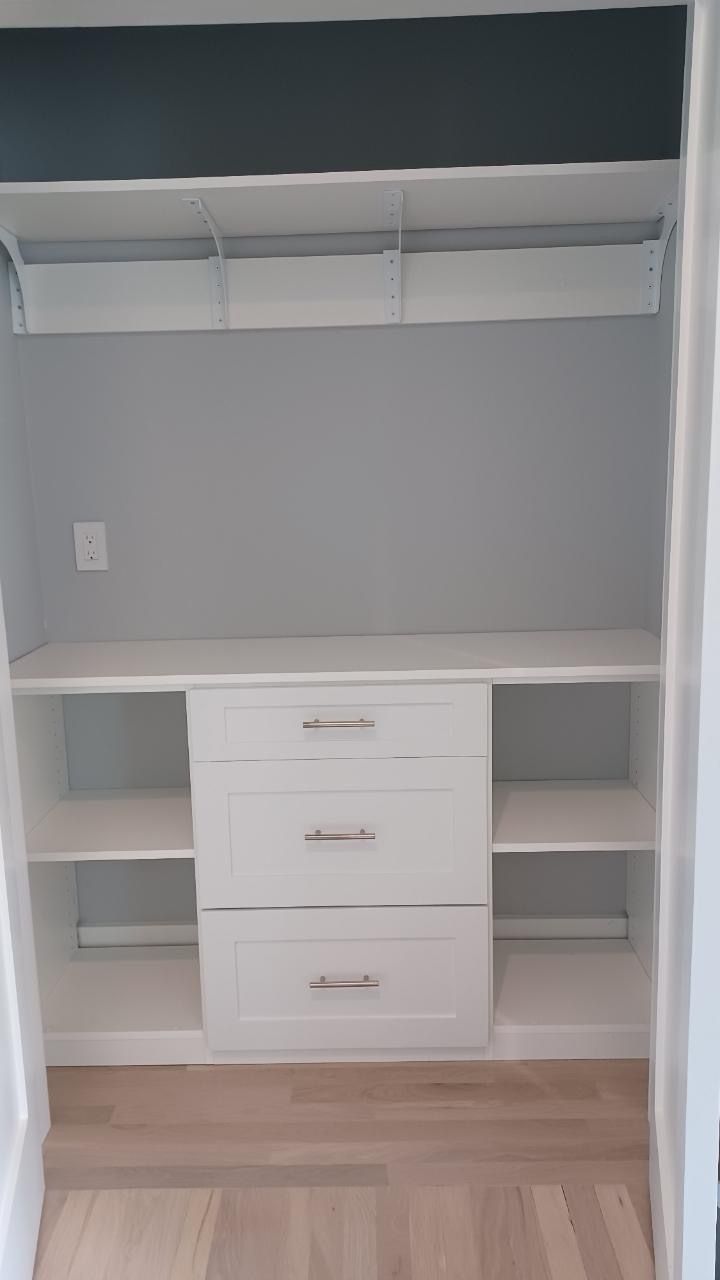 White closet interior with shelves, drawers, and hanging rod, against gray walls.