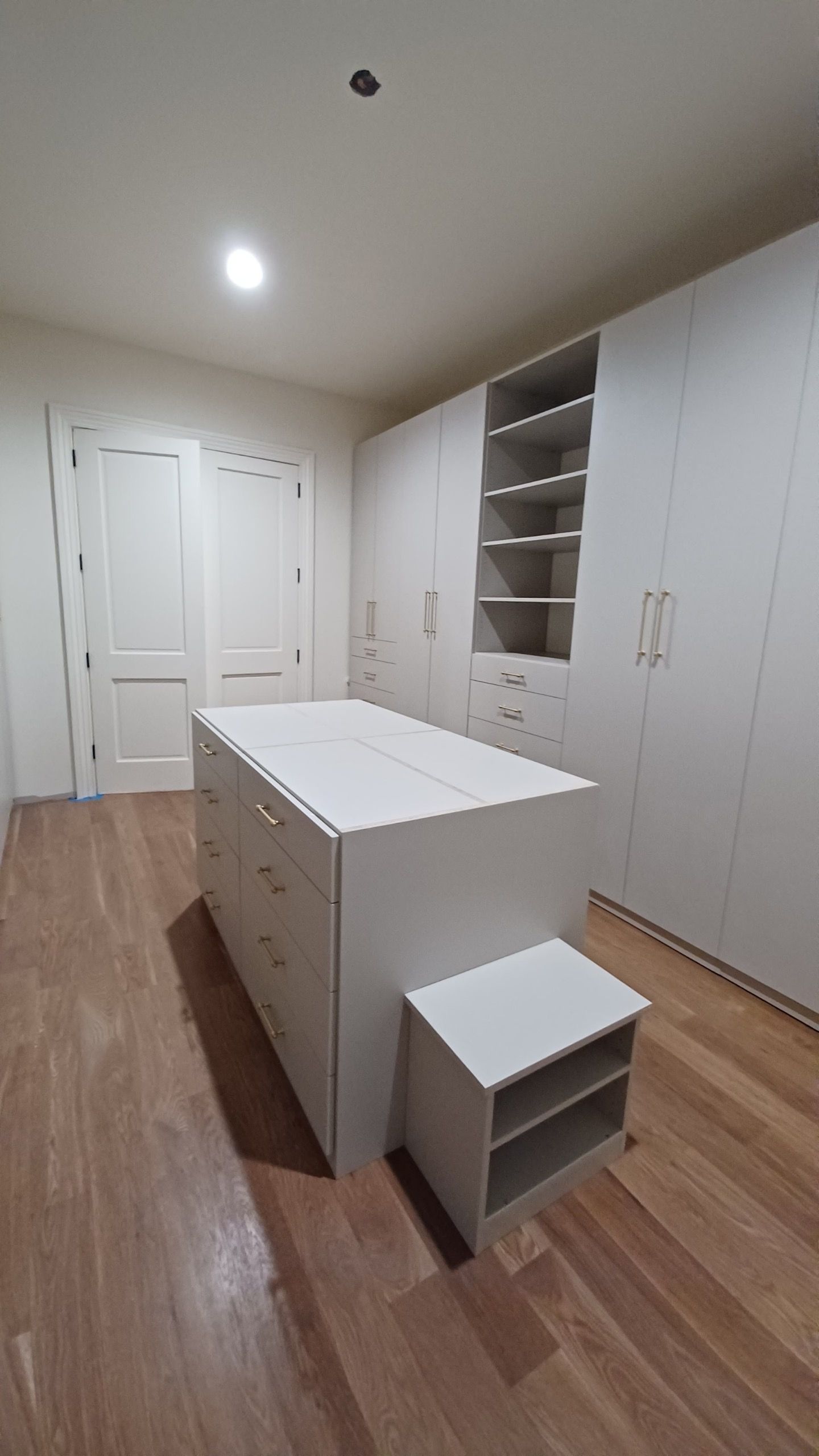 White walk-in closet with a center island, built-in shelves, and wooden floors. Includes a small bench.