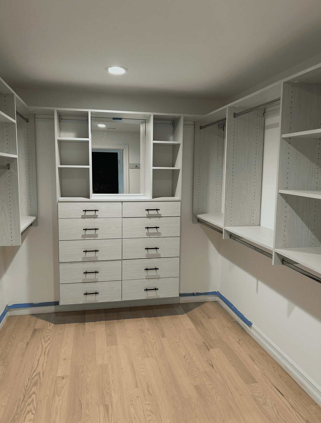 Walk-in closet with white shelves and drawers, wooden floor. Center: mirror and drawers.