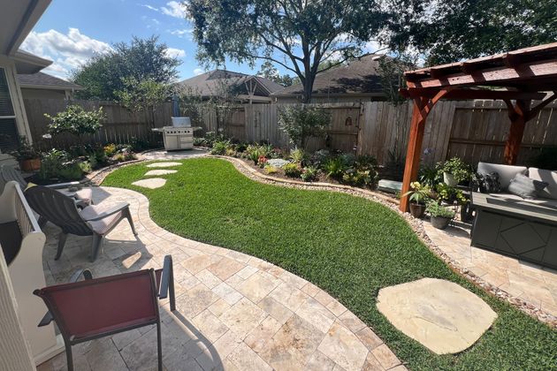 Sunny backyard patio with lawn, stone path, chairs, pergola, and garden beds