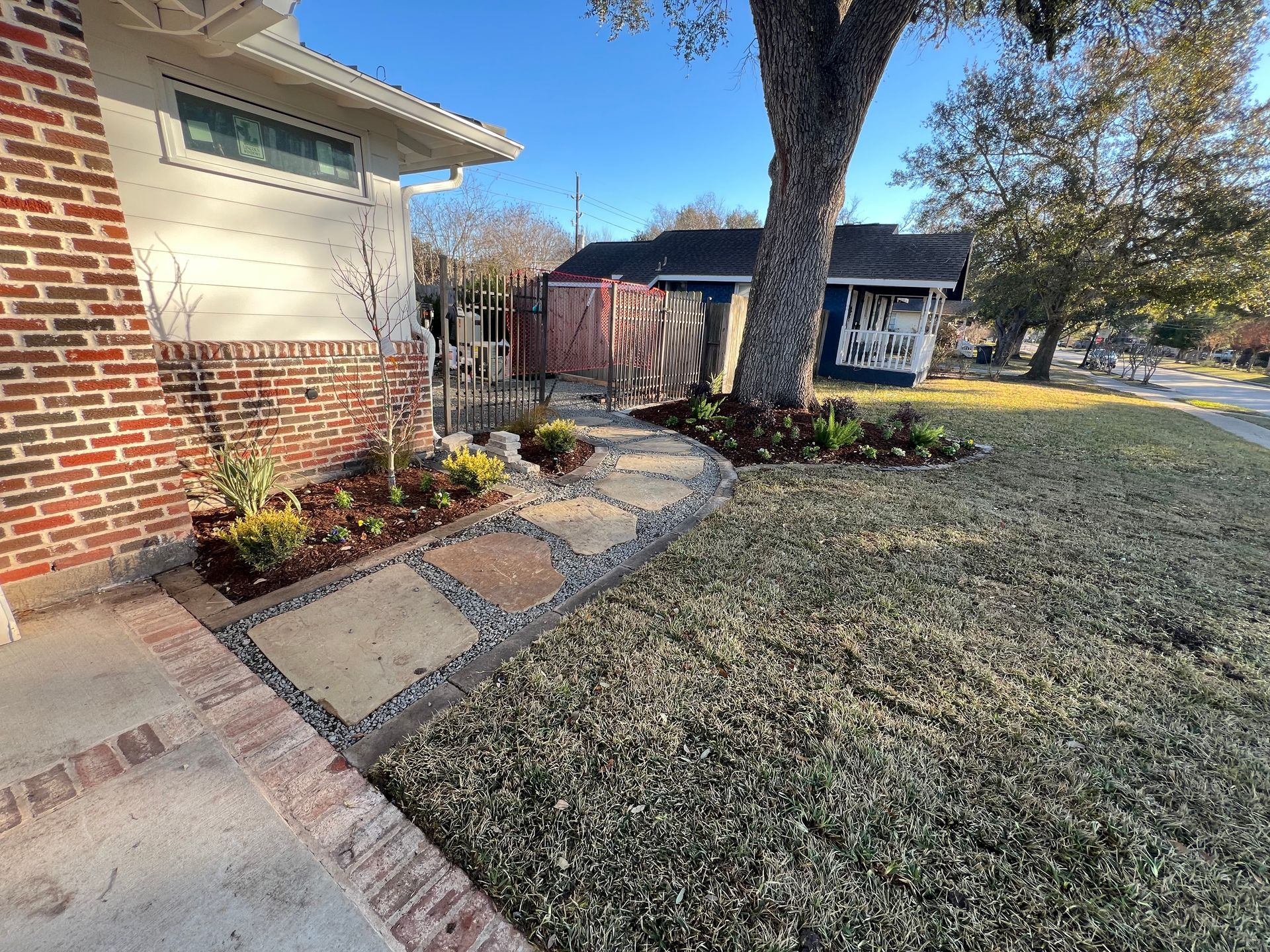 Front yard walkway beside brick house, with flower beds, tree, and small shed under a clear sky.