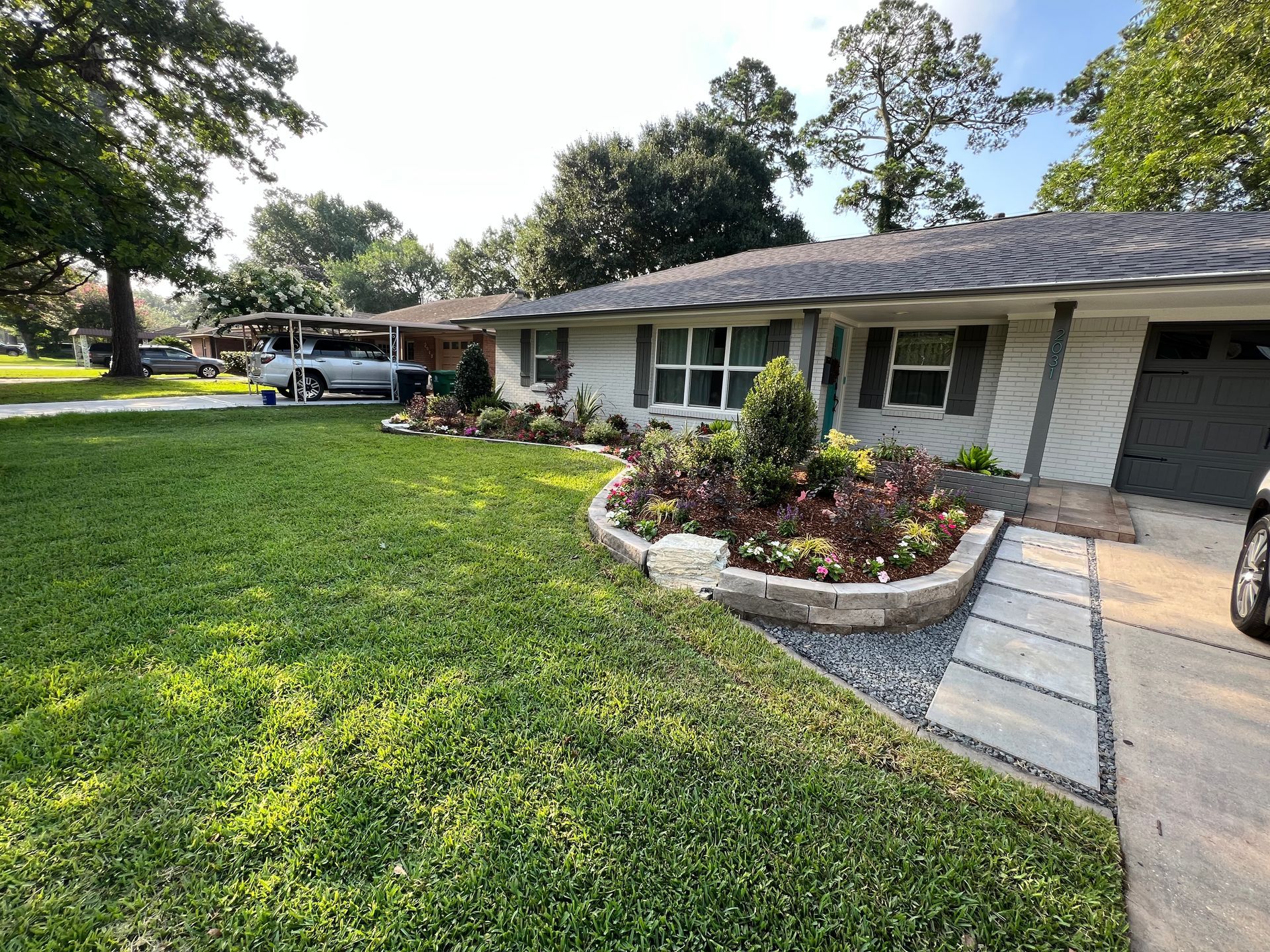 Single-story suburban house with a green lawn, flower beds, and a concrete walkway under a bright sky