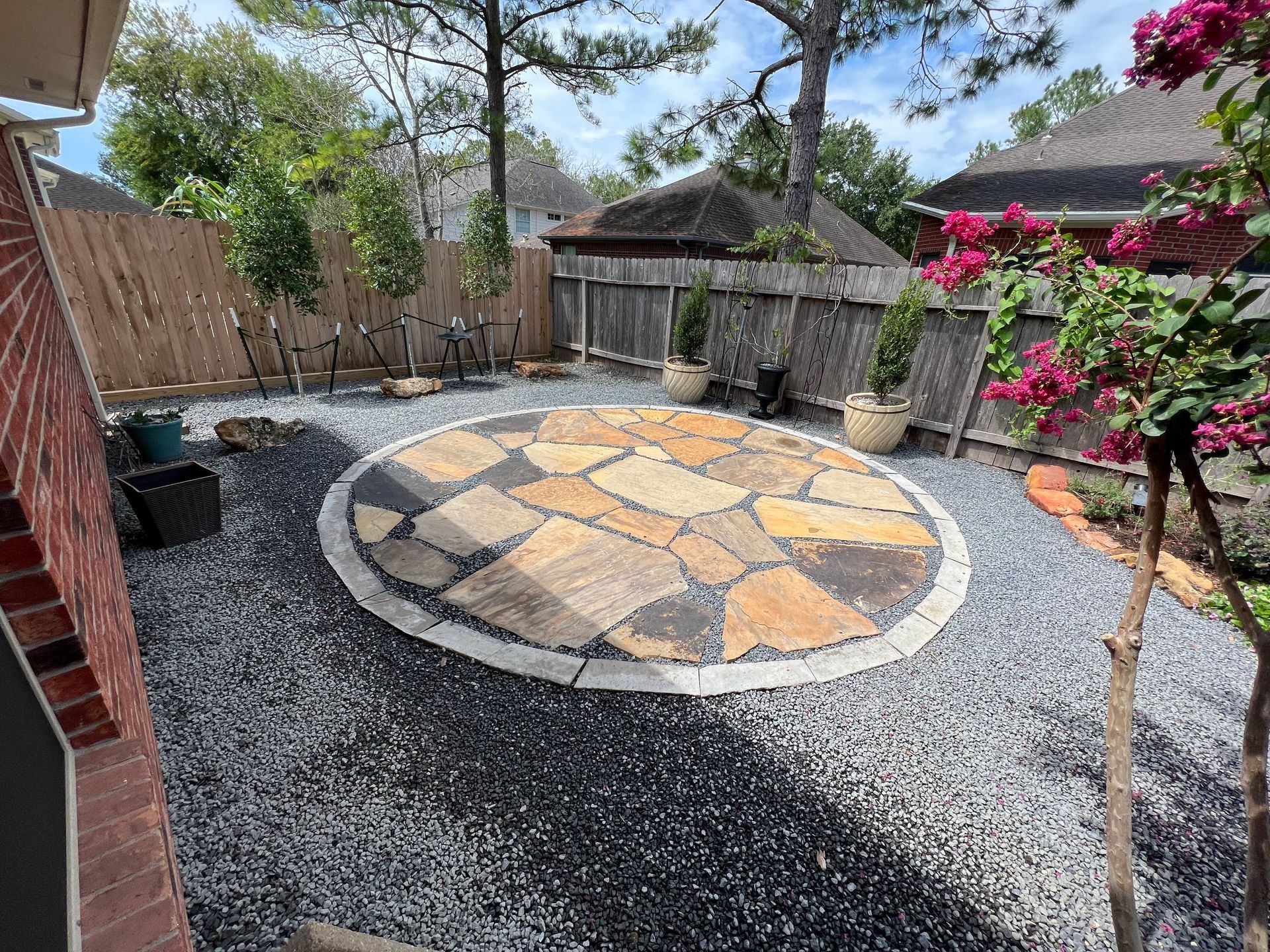 Backyard patio with circular stone paving, gravel ground, and a flowering bougainvillea near a fence.