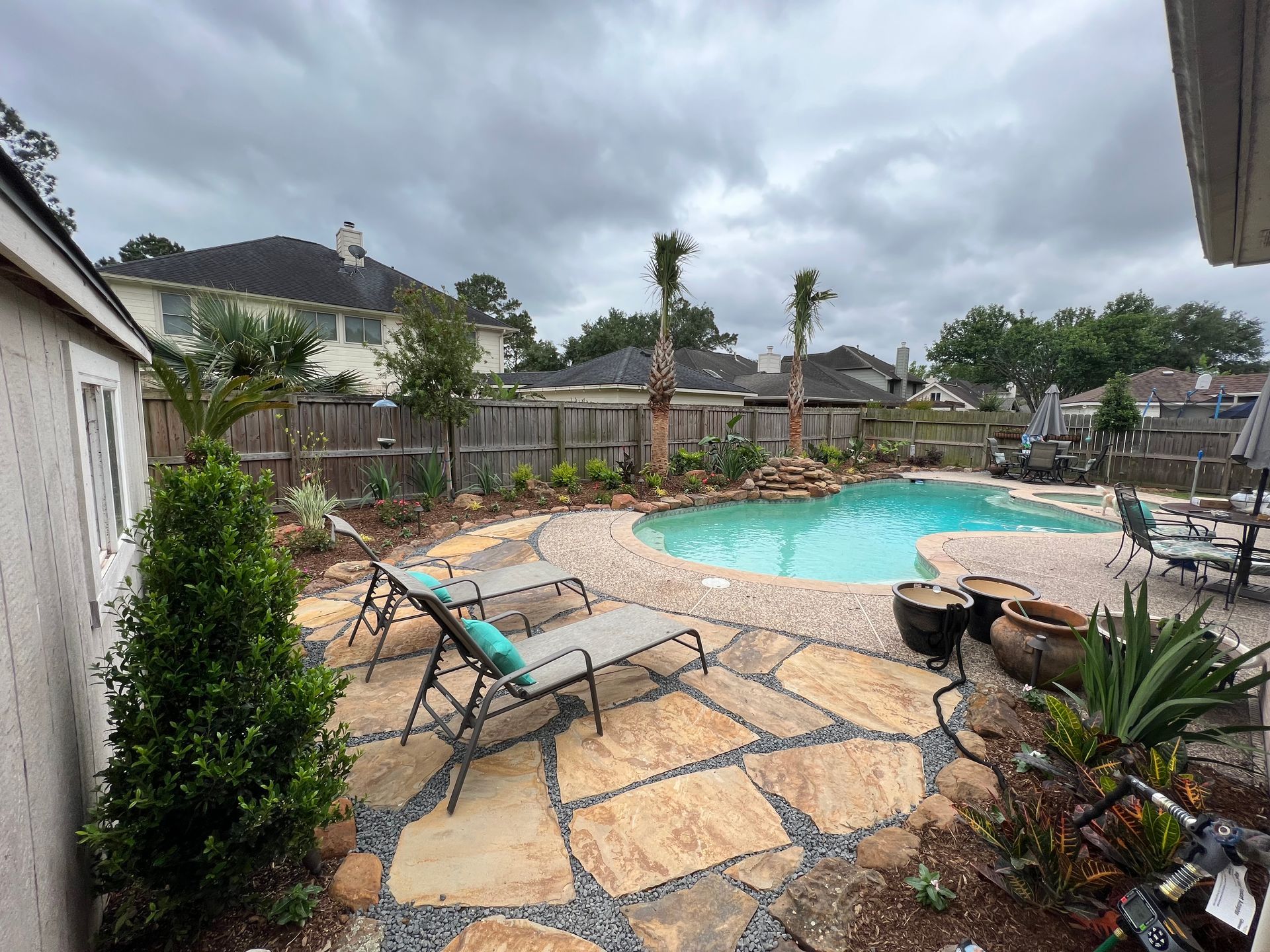 Backyard patio with stone paving, lounge chairs, and a turquoise swimming pool under cloudy skies