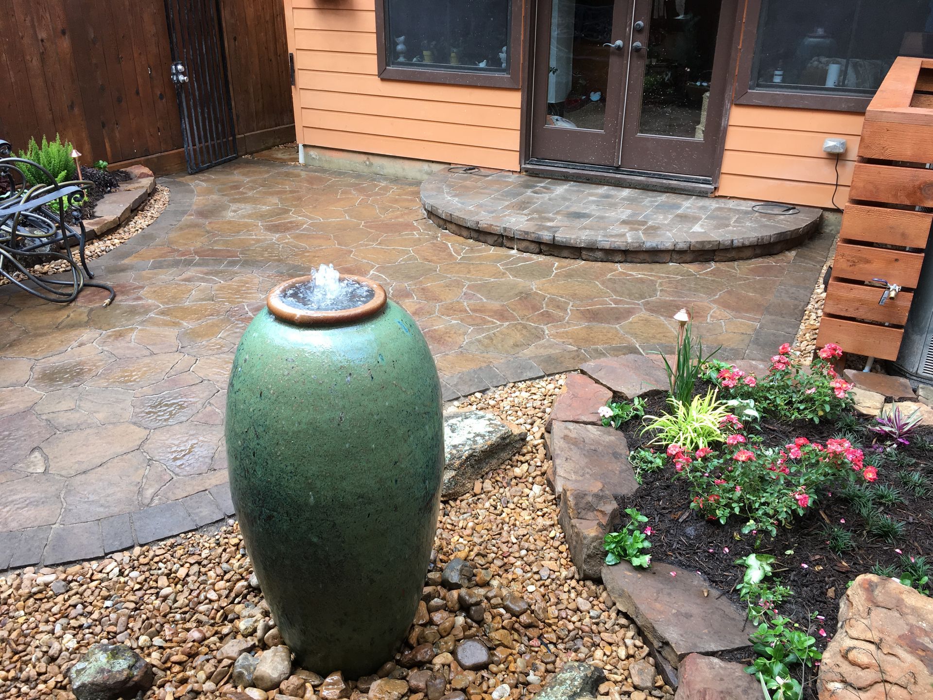 Green ceramic garden urn fountain in a rock bed outside a house, with wet stone patio and flowers nearby