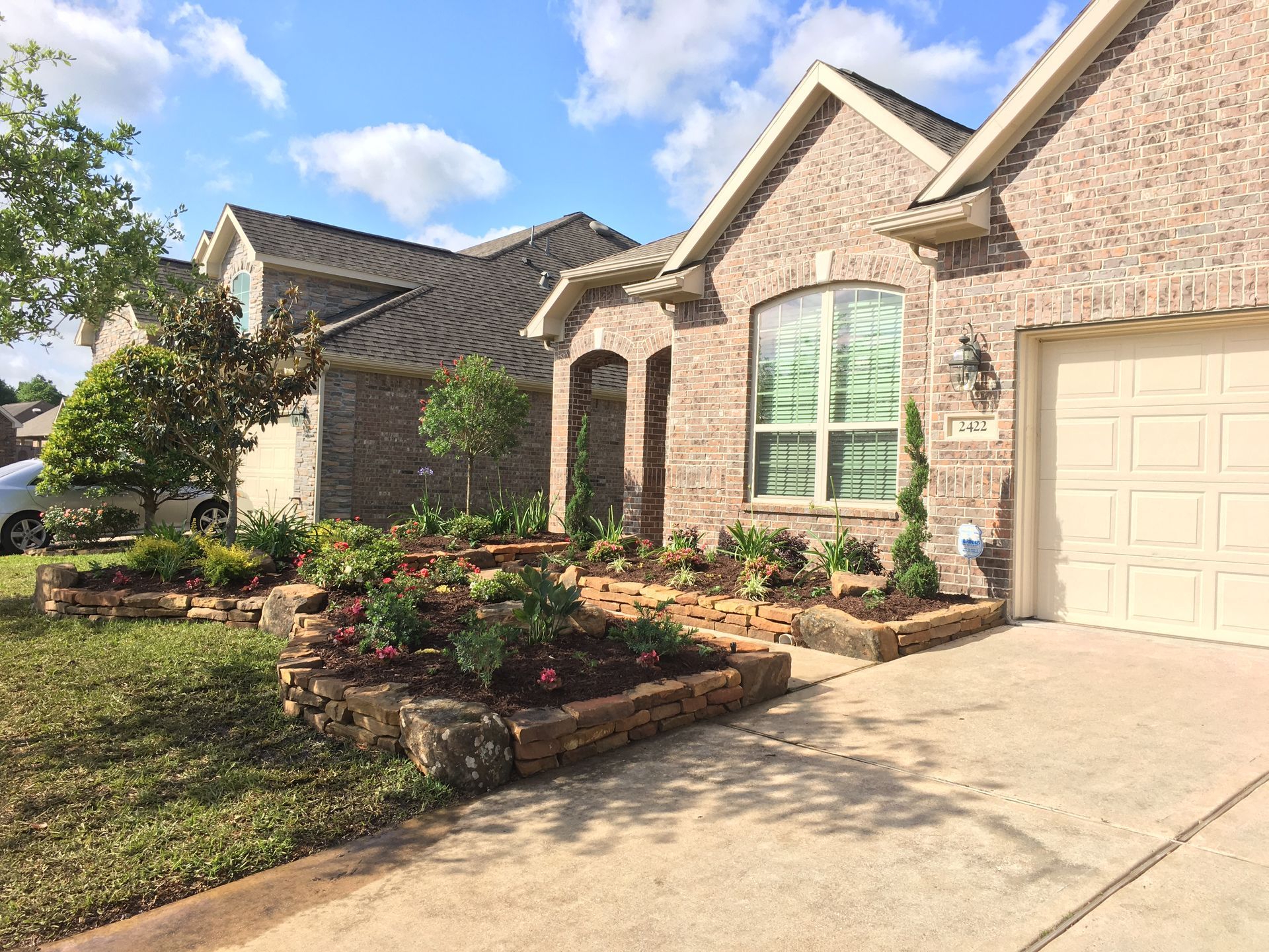 Brick house exterior with driveway, landscaped garden beds, and a garage under a blue sky