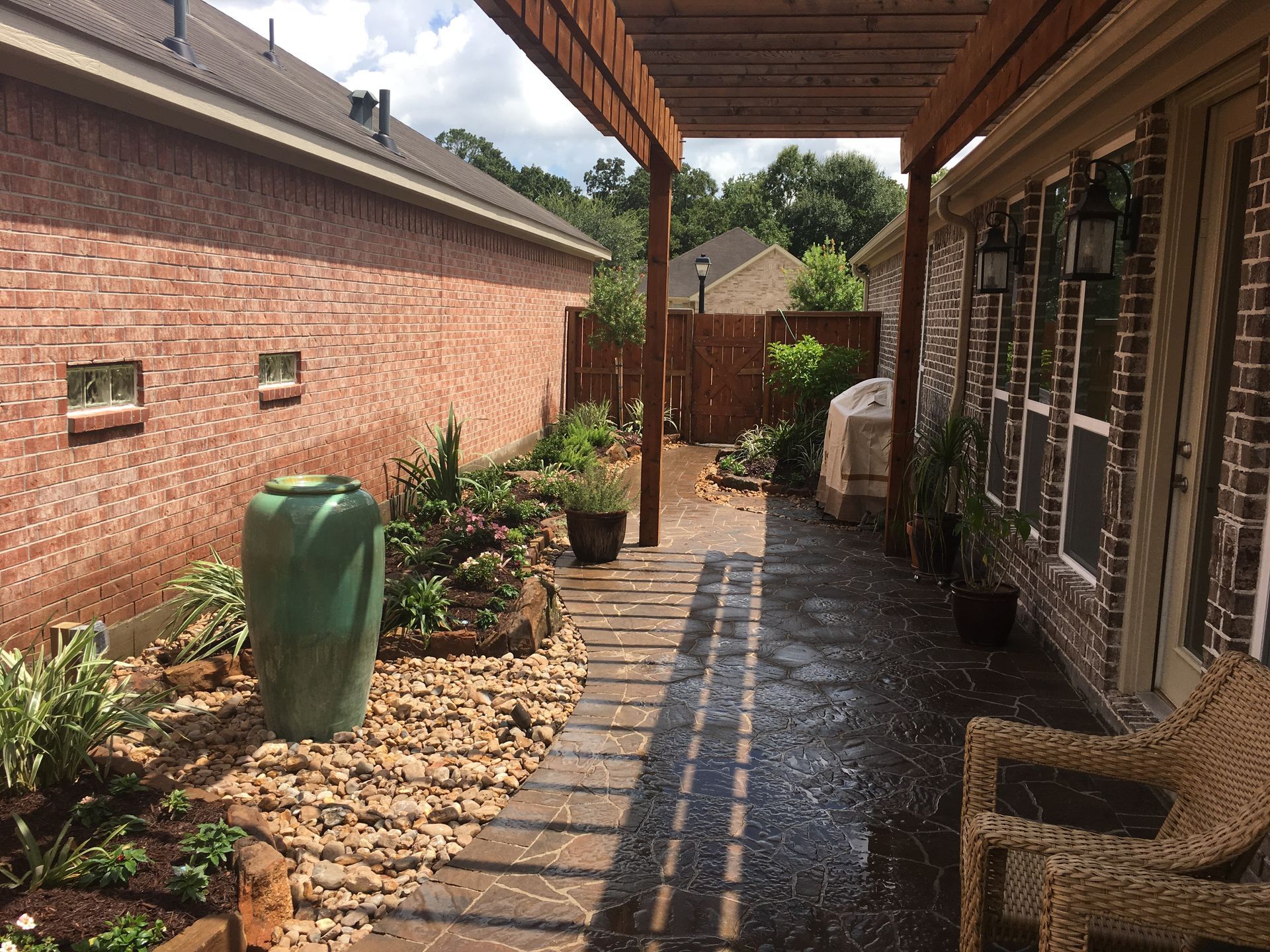 Covered brick patio walkway beside a house, with wet floor, potted plants, and a wicker chair