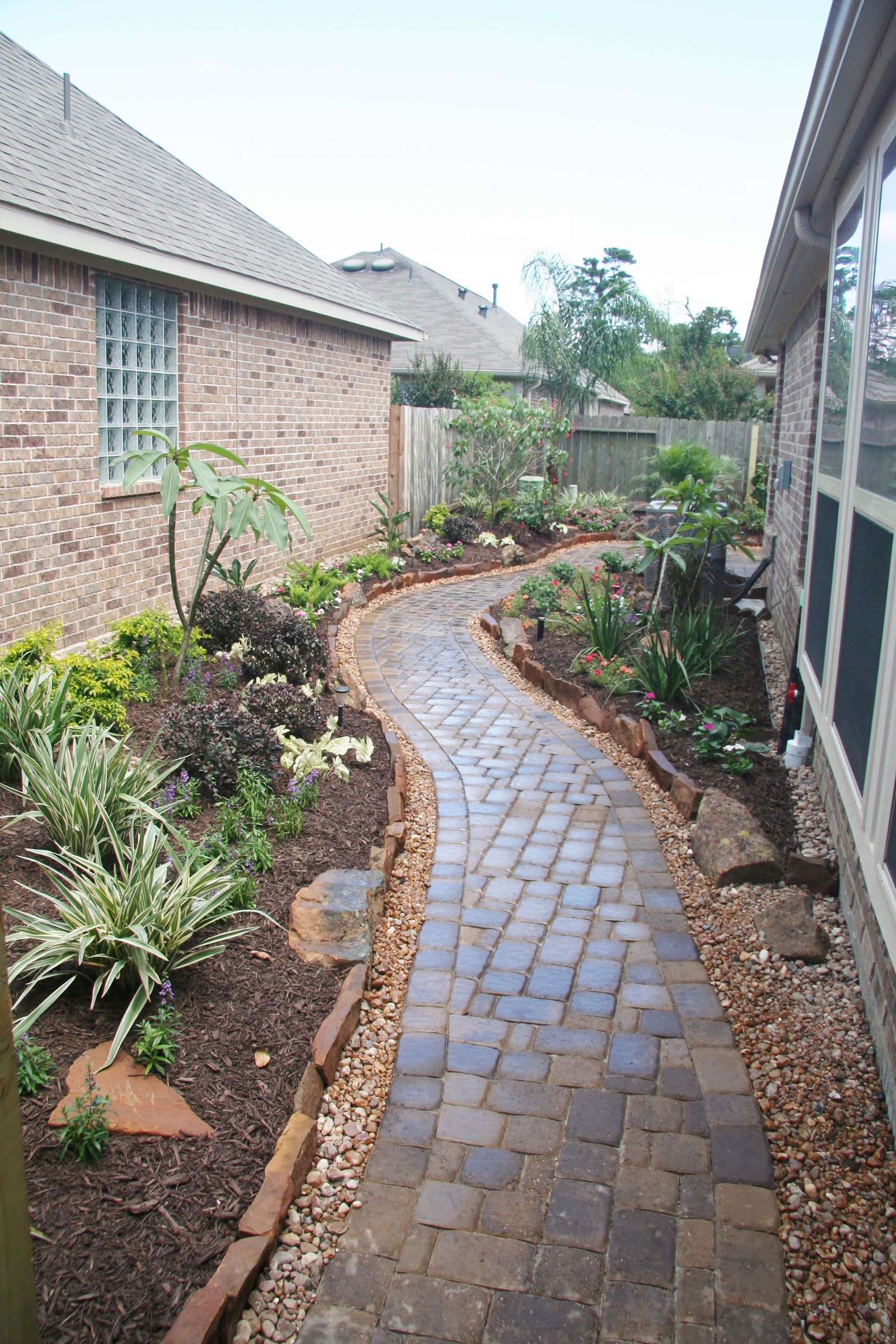 Curved brick walkway through a landscaped side yard beside a brick house and fence, bordered by mulch and plants