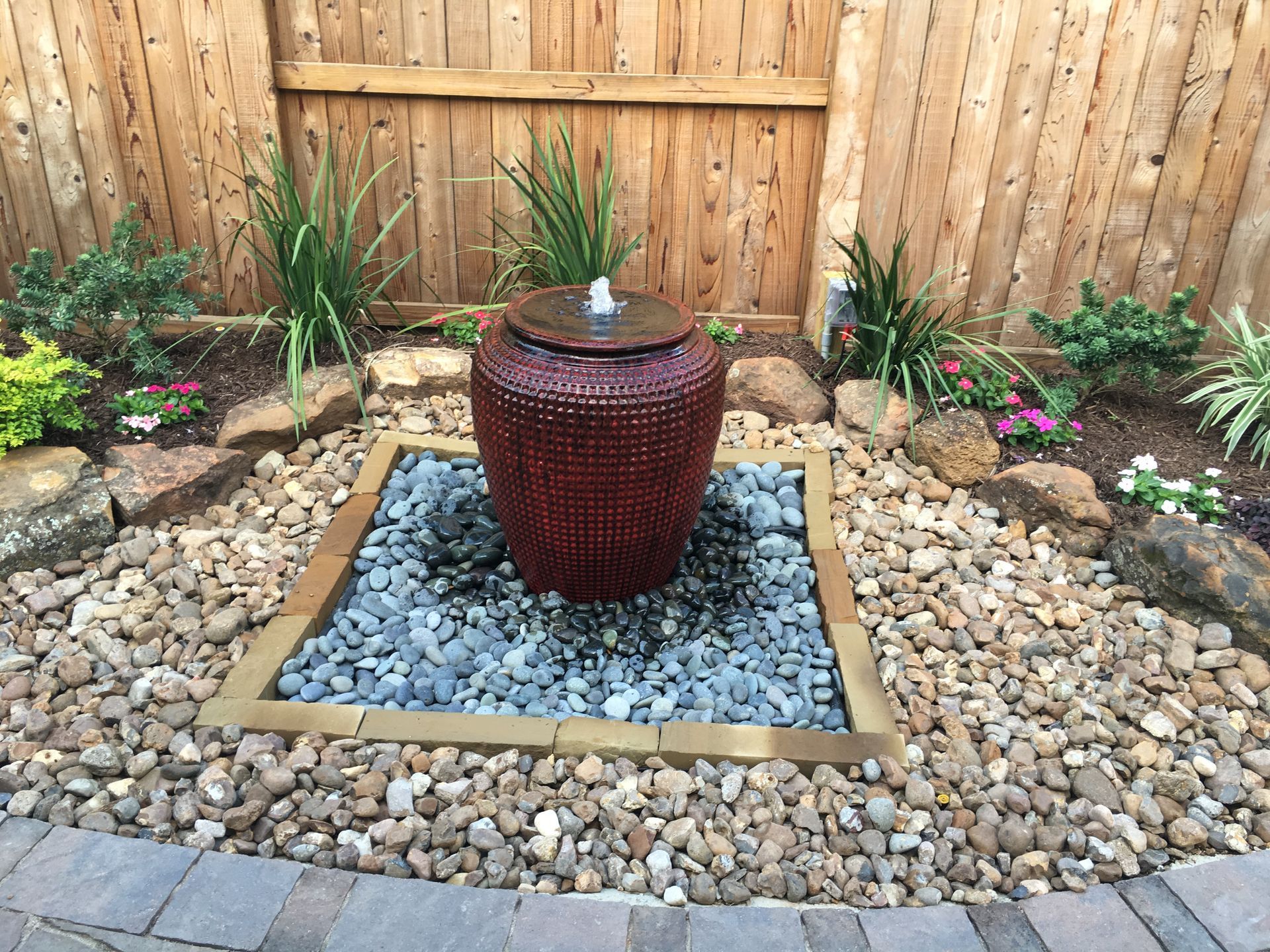 Backyard garden fountain with red urn centerpiece, pebbles, stone border, and wooden fence backdrop