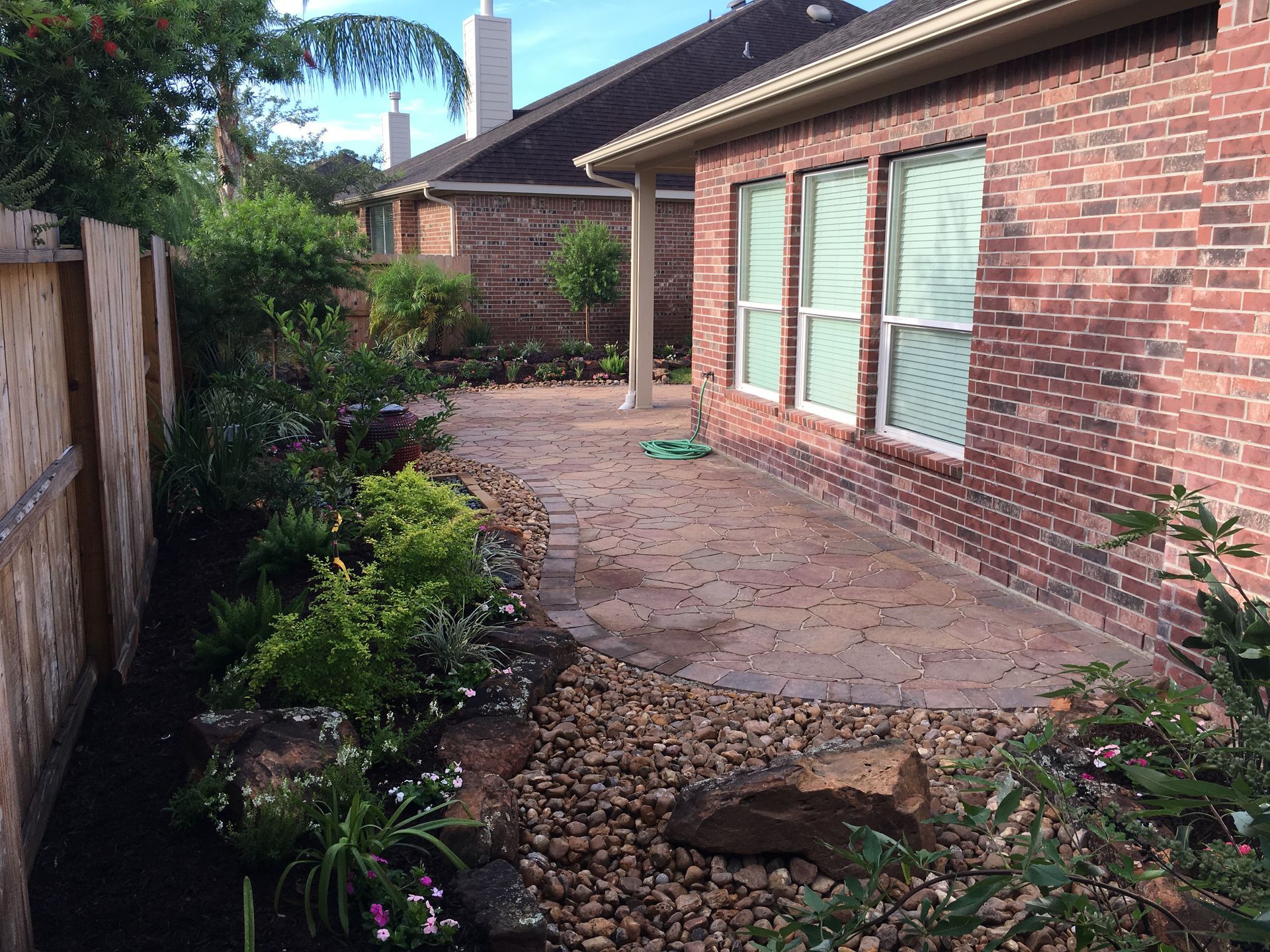 Brick patio beside a brick house, with a narrow garden path and flower beds along a wooden fence.