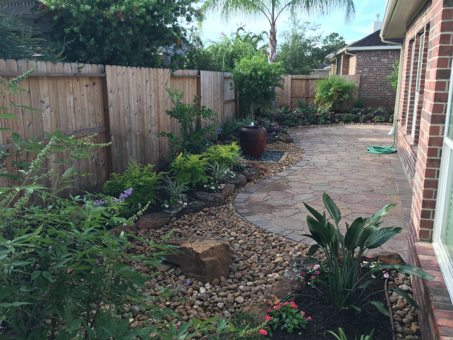 Narrow backyard garden path with stone pavers, gravel borders, wooden fence, and brick house wall