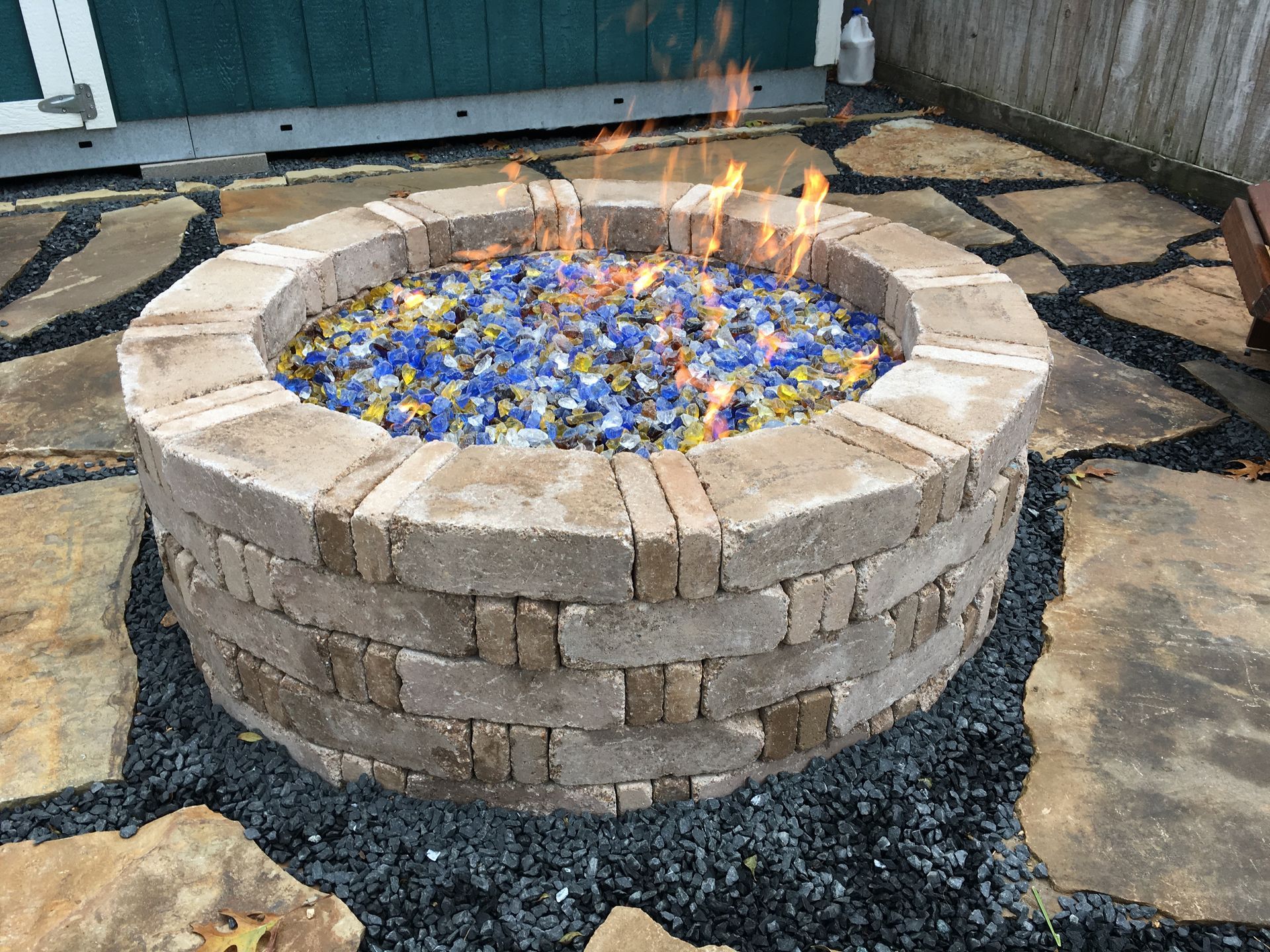 Stone fire pit with orange flames over blue glass on a patio, surrounded by pavers and gravel