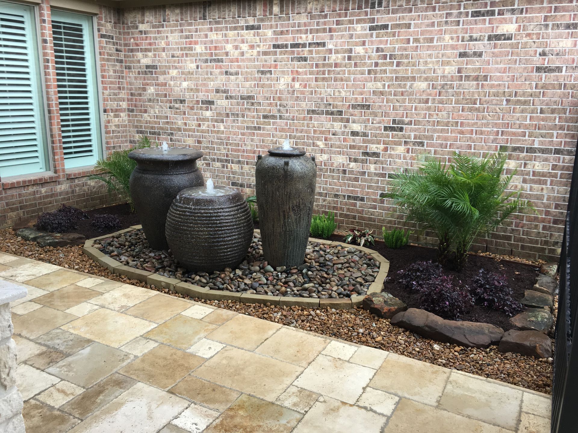 Brick courtyard with three decorative black urns and a small green shrub bed beside a brick wall.