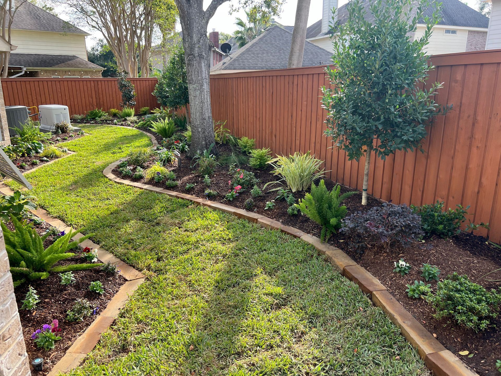 Curved green backyard path bordered by wood edging, mulch beds, shrubs, and a wooden fence