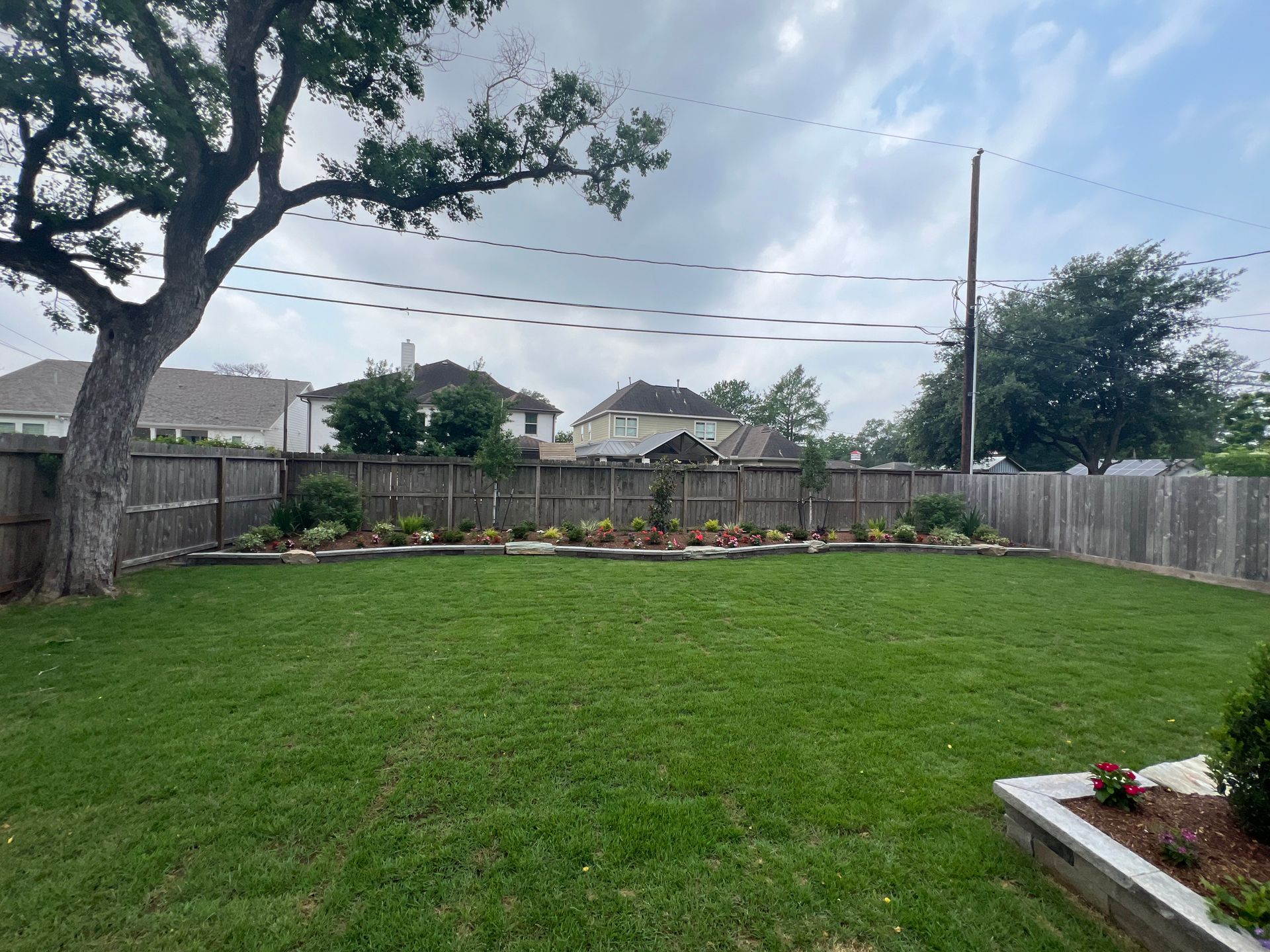 Backyard with green lawn, wooden fence, trees, and a flower bed along the back wall