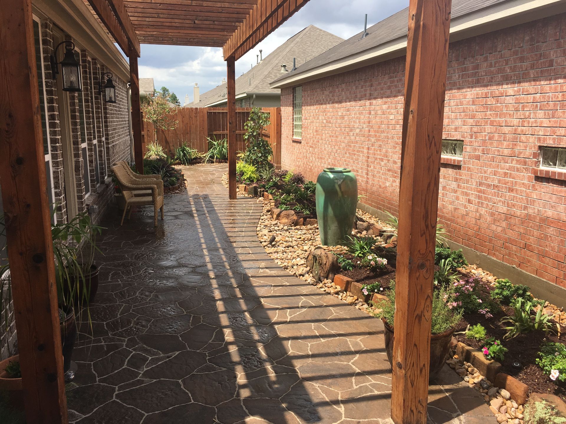 Narrow backyard patio with brick walls, wooden pergola, potted plants, and a stone walkway in sunlight