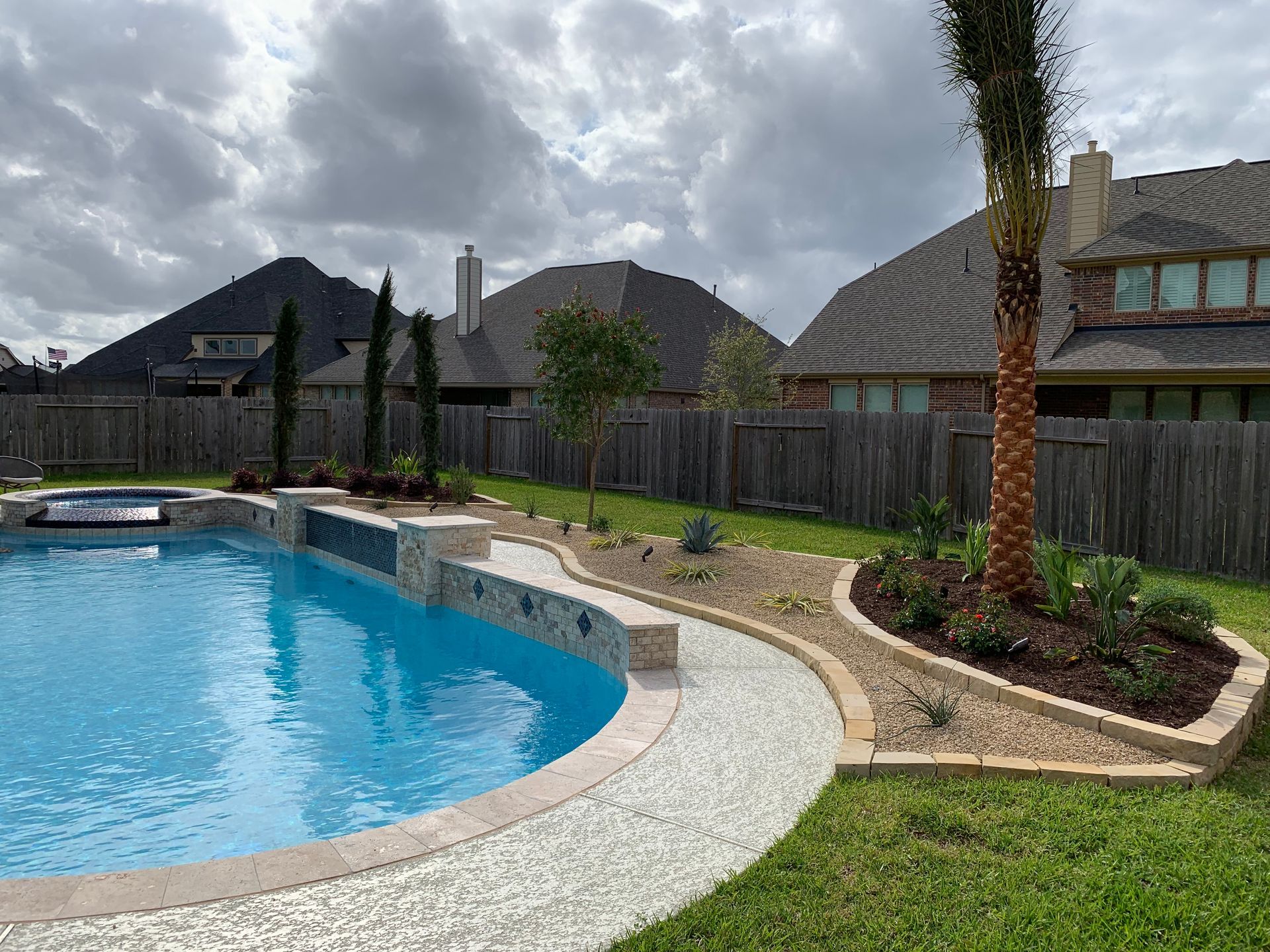 Backyard pool with curved stone path, lawn, fence, and cloudy sky beside modern houses