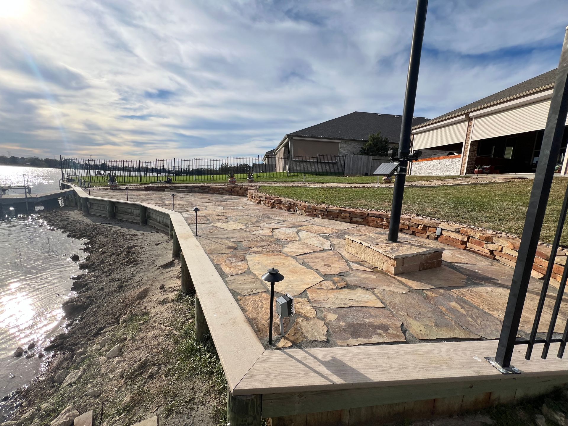 Stone patio beside a shoreline with black lamp posts and a building under a cloudy sky