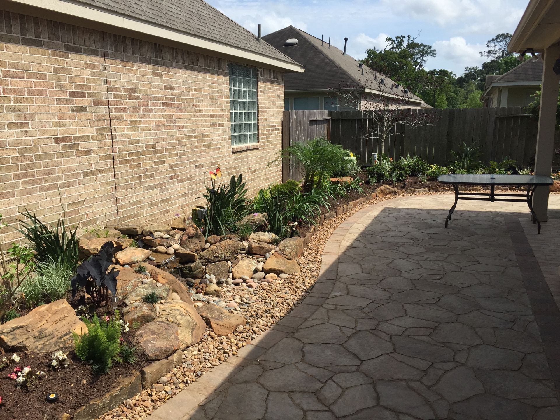 Stone garden path beside brick house and flower bed, with a patio table in a sunny backyard.