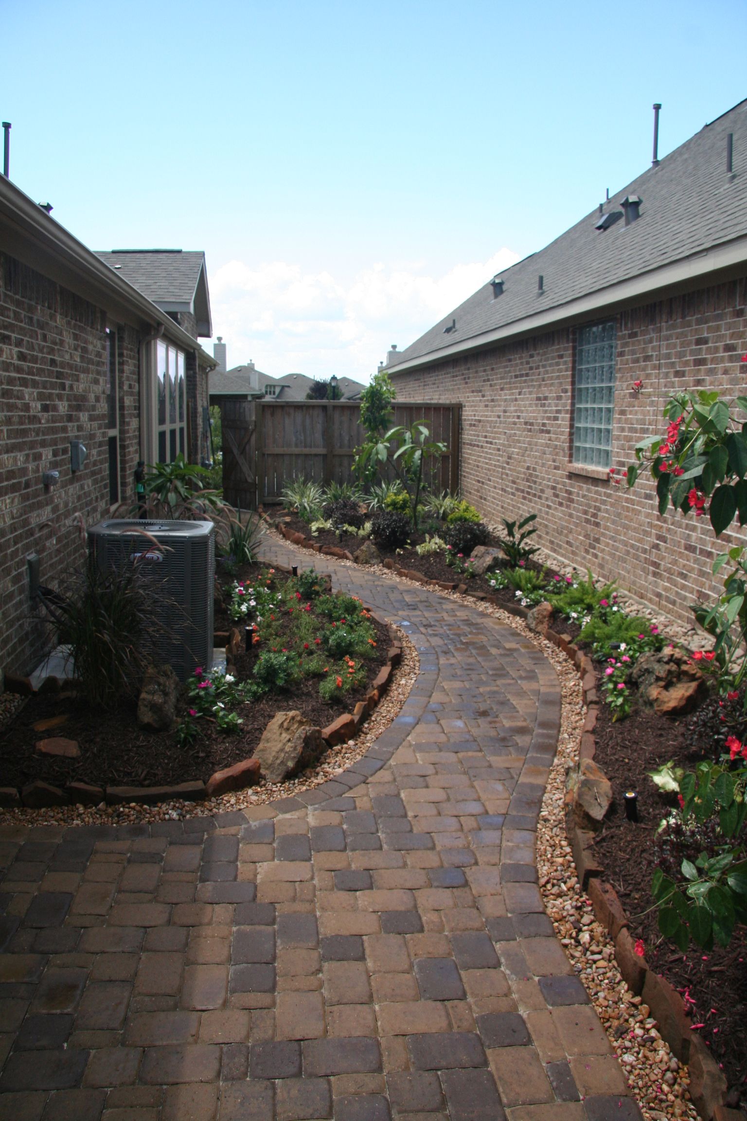 Curved brick walkway between two houses, lined with small plants and flowers under a bright sky
