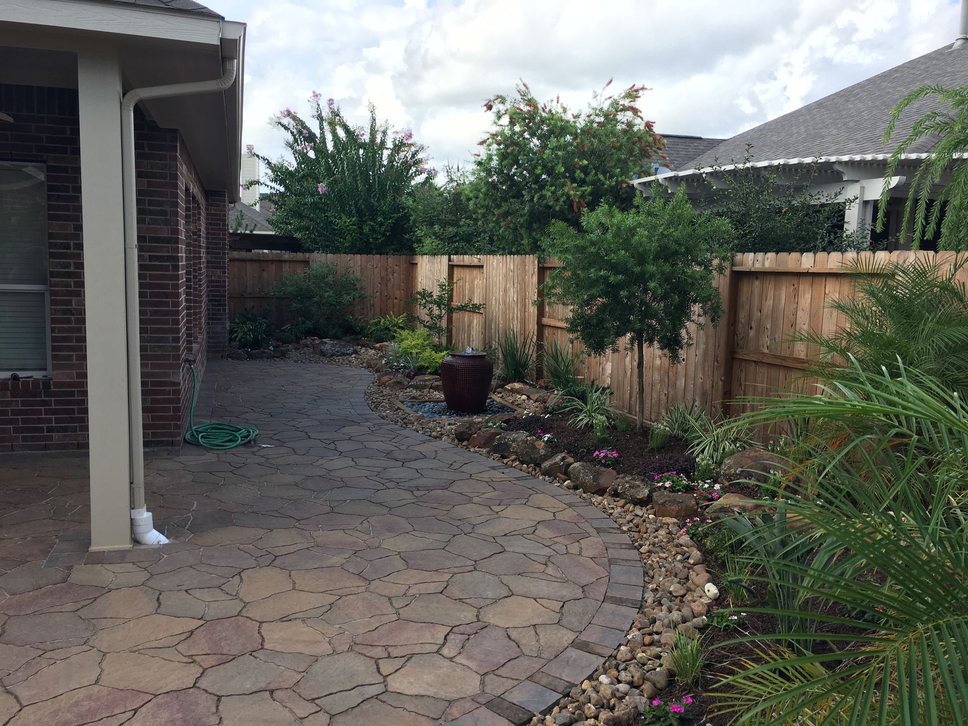 Backyard patio with stone pavers, wooden fence, and shrubs beside a brick house