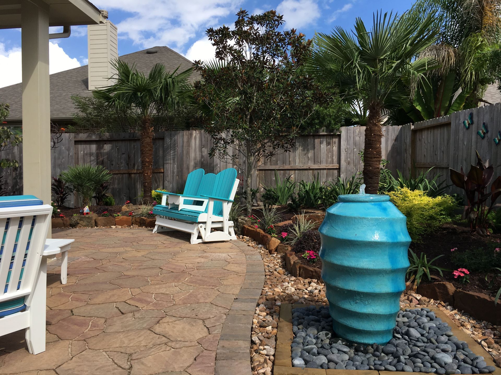 Patio with turquoise chairs, a blue ceramic vase, stone pavers, and a fenced garden