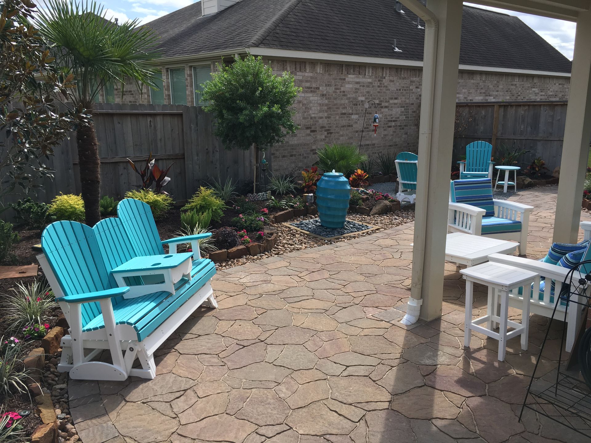 Backyard patio with turquoise Adirondack chairs, a white table, and potted plants by a brick wall.