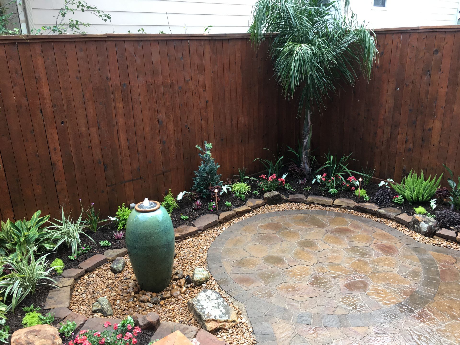 Small backyard patio with circular stone pad, wooden fence, potted planter, and surrounding greenery