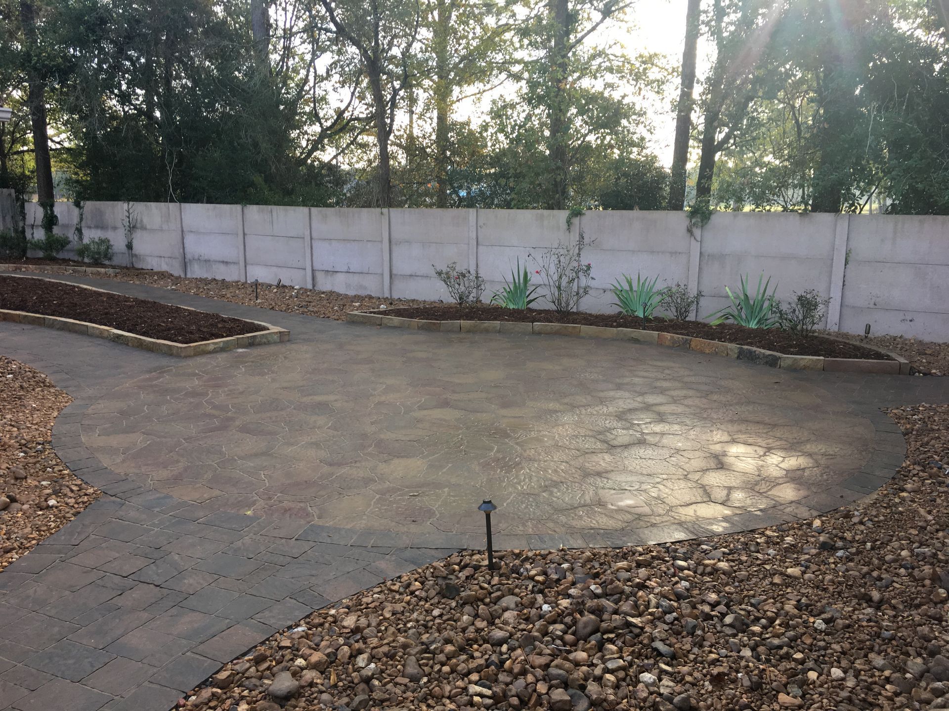 Empty landscaped courtyard with stone paving, white wall, and trees in the background.