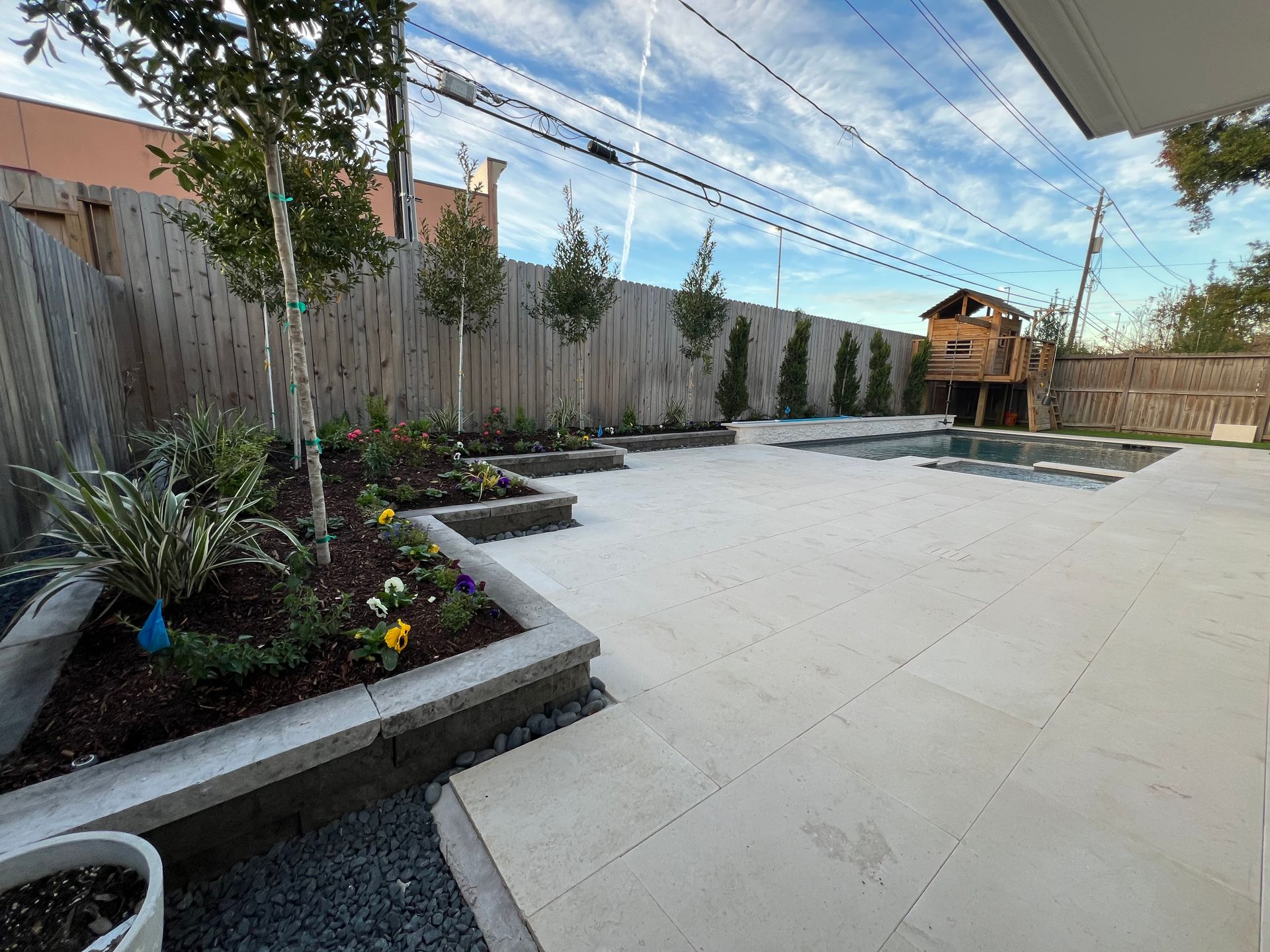 Backyard patio with white pavers, raised planters, trees, and a wooden fence under a shade canopy