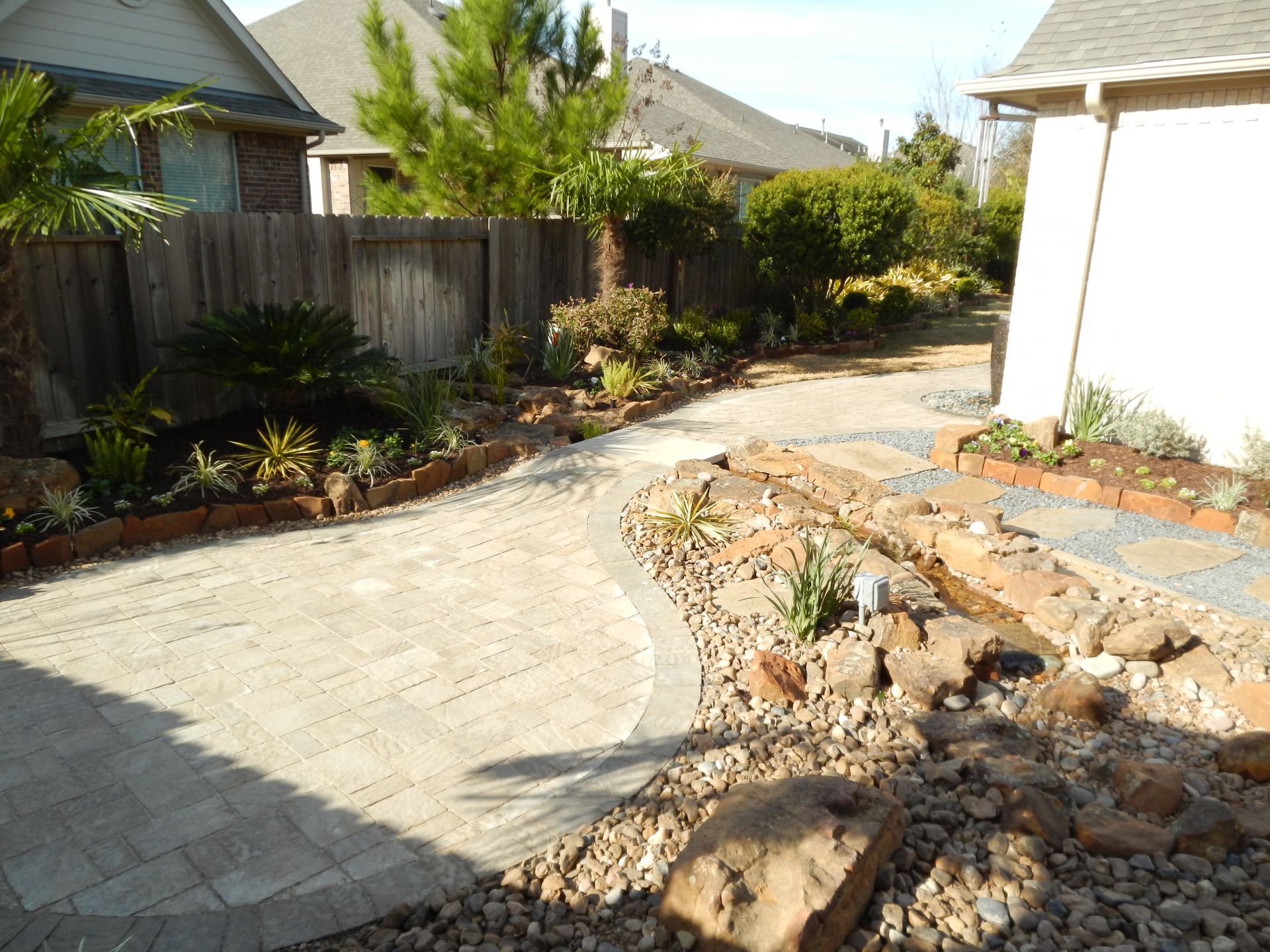 Curved stone walkway beside a white house, with garden beds, rocks, shrubs, and a wooden fence.