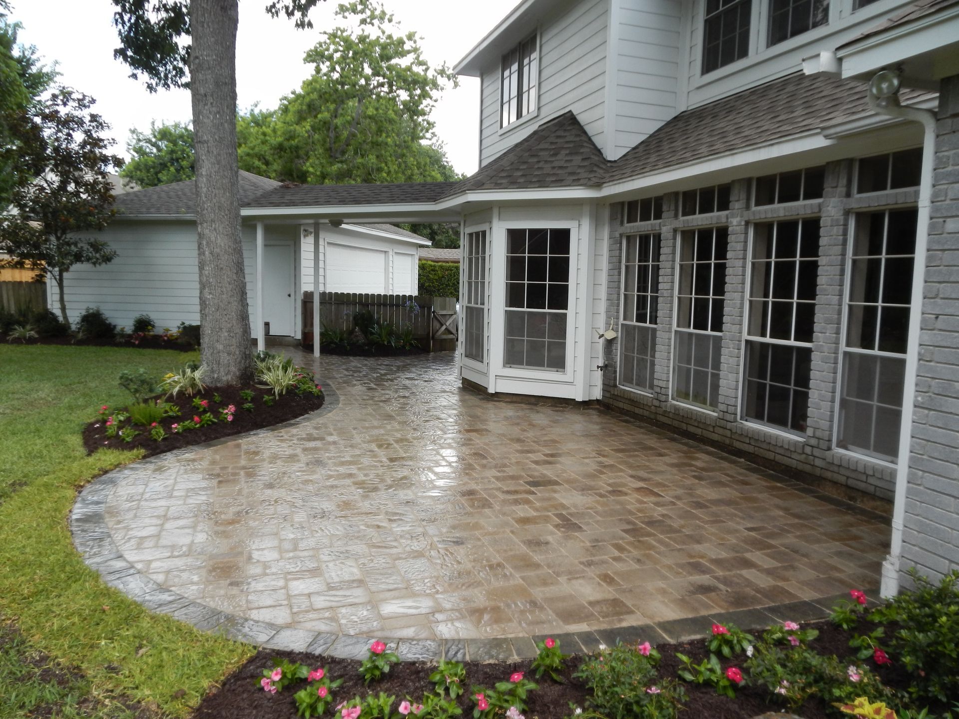 Patio with light stone pavers beside a gray house, bordered by flower beds and green lawn.