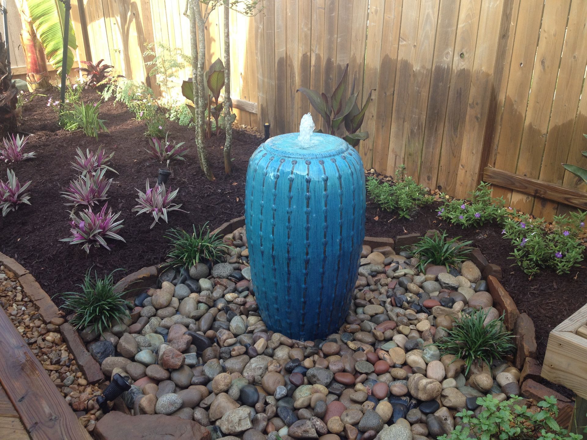 Blue ceramic garden fountain on a bed of river stones, surrounded by plants and a wooden fence.