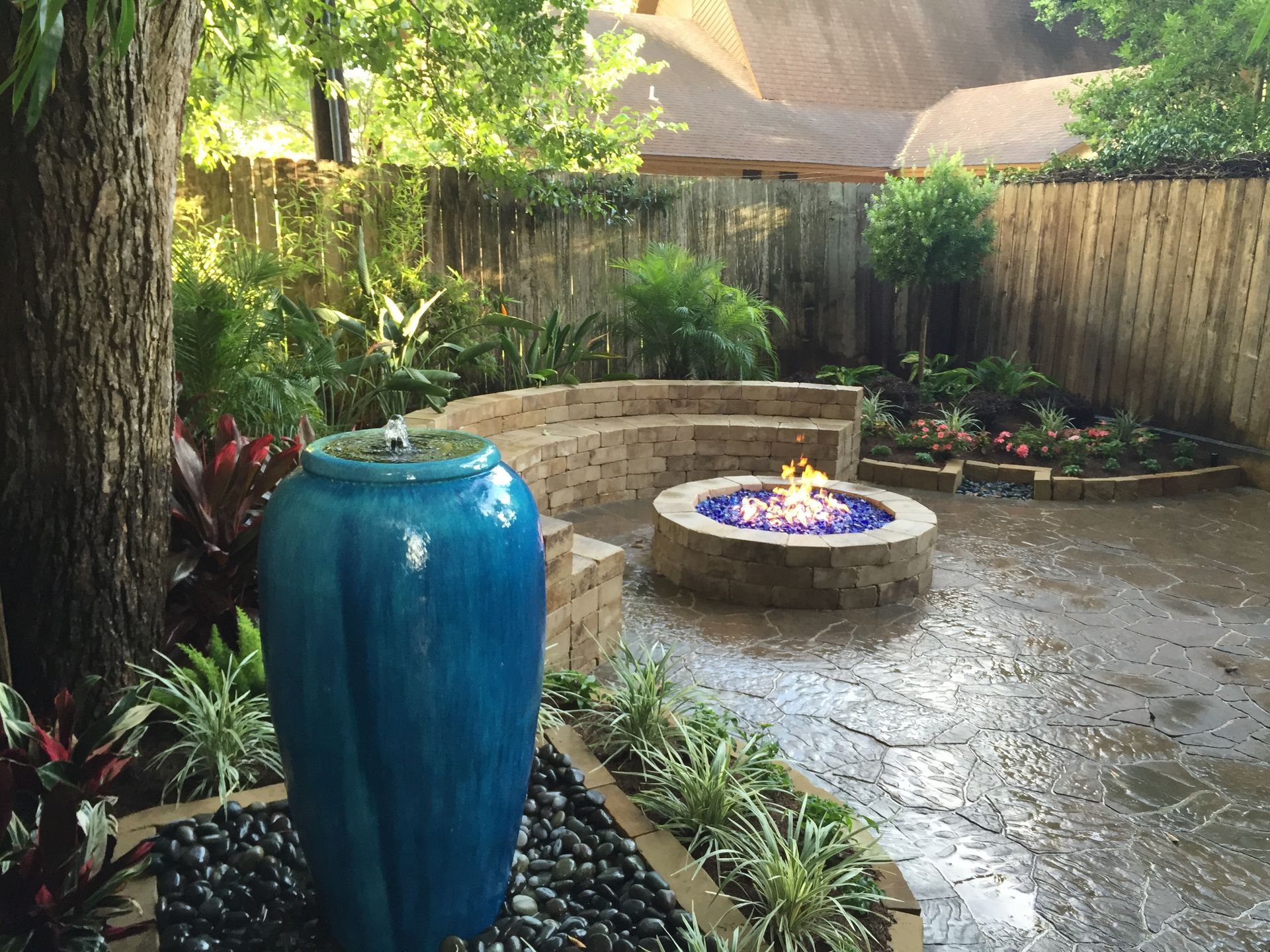 Small garden patio with a blue ceramic vase, circular fire pit, and lush plants along a fence.